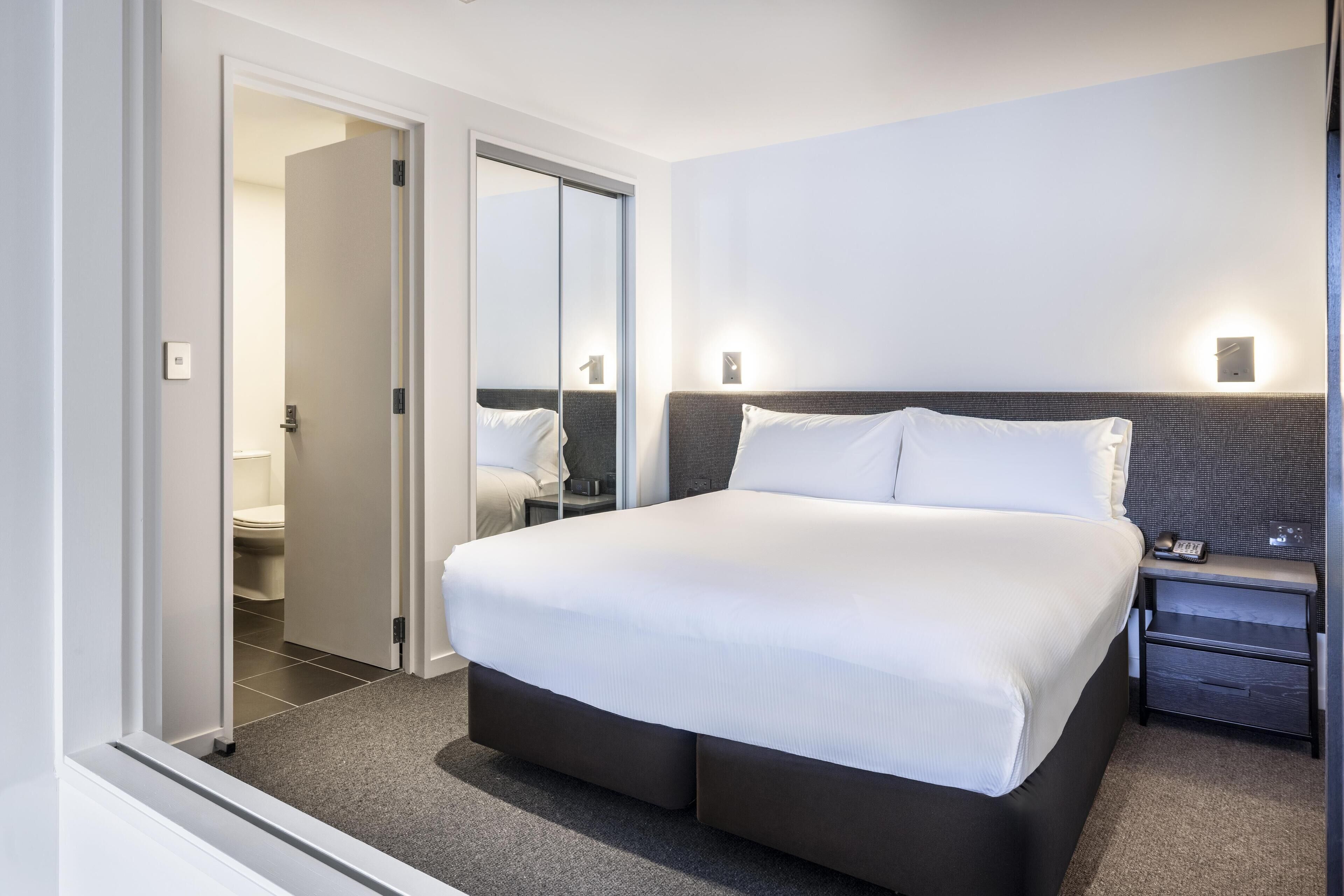 Moderni hotellihuone, jossa on suuri sänky, yöpöytä, yövalaisin ja kylpyhuone, joka näkyy avoimesta ovesta.