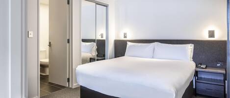 A modern hotel room with a large bed, a bedside table, a nightstand, and a bathroom visible through an open door.