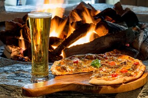 A pizza on a wooden board with a glass of beer in front of a fire. - Hilton Queenstown Resort & Spa (Kawarau Falls)