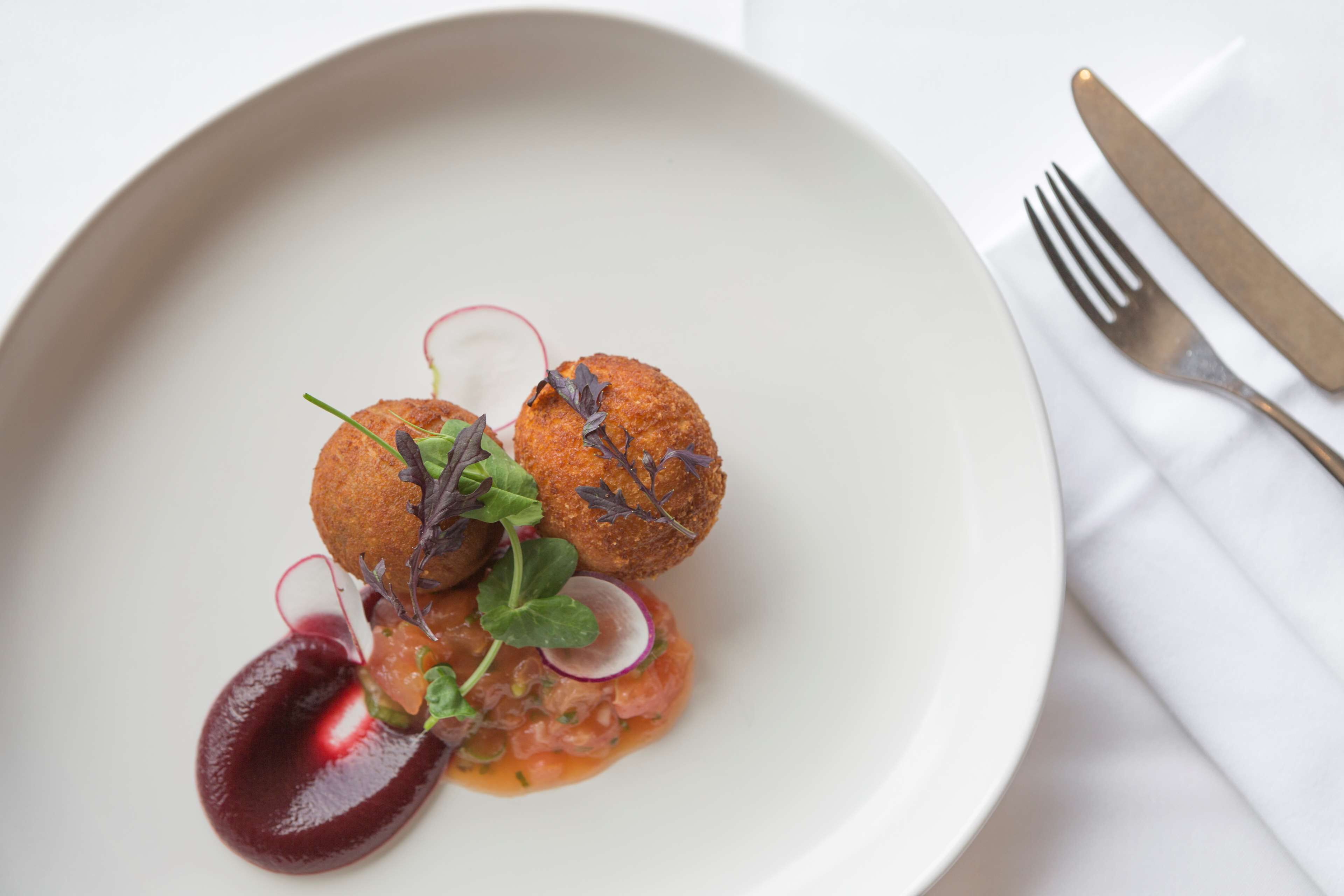A plate of food with two round, golden-brown items, possibly croquettes, garnished with greens and radish slices, and a dark sauce.