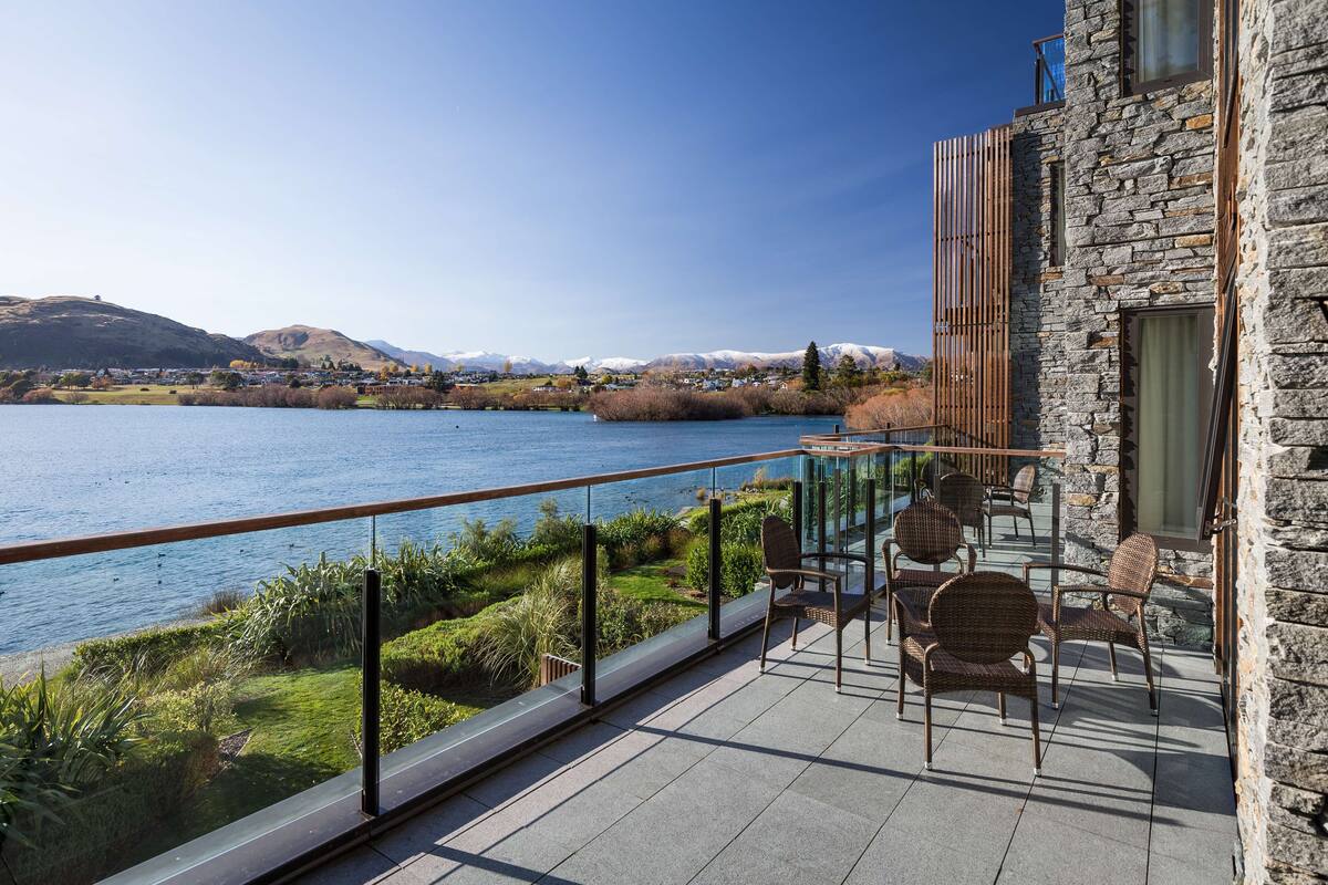 A balcony with a view of a lake and mountains, furnished with wicker chairs and a glass railing.