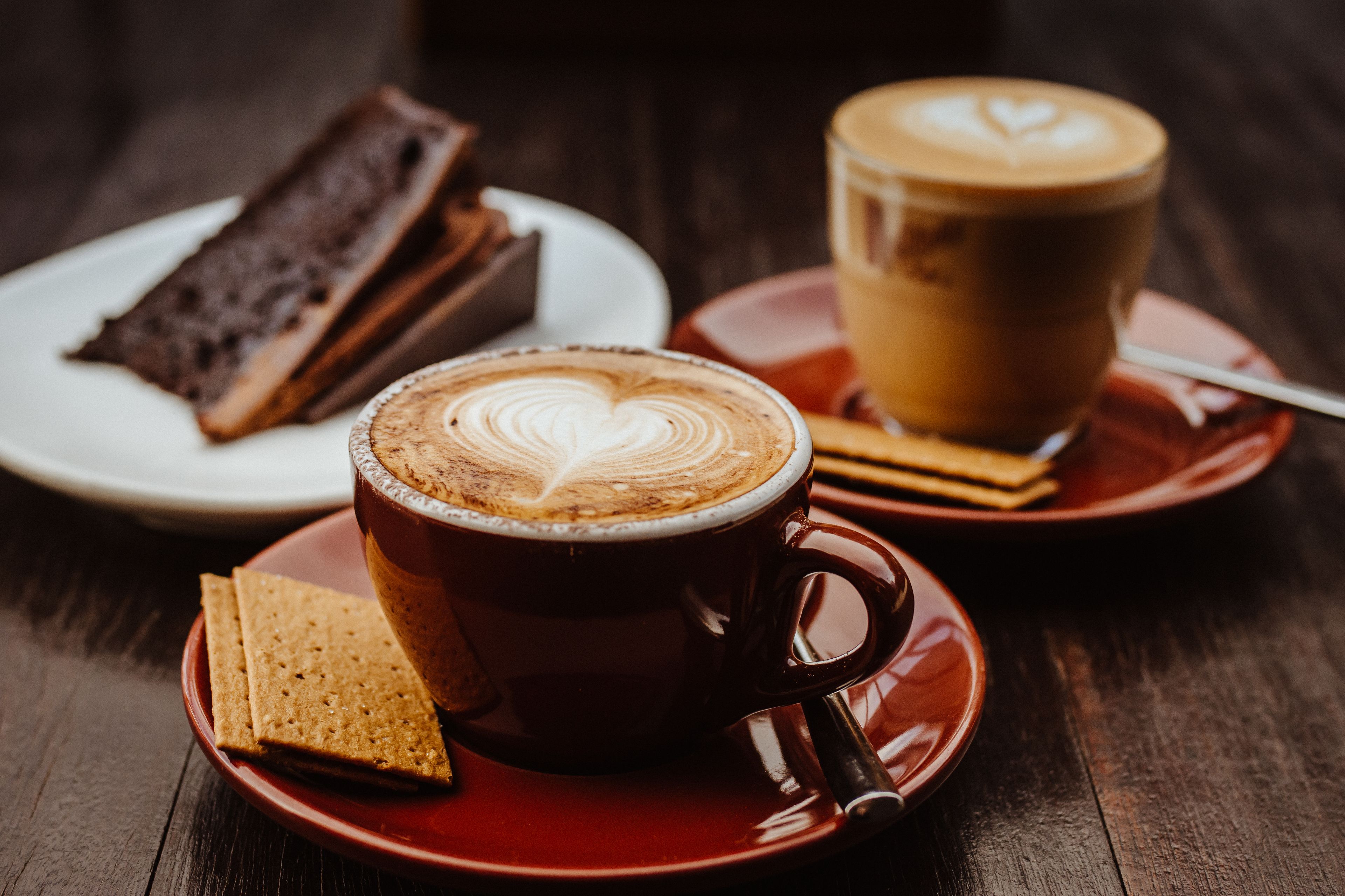 A cup of coffee with latte art on a saucer, a plate with a slice of cake, and a plate with a biscuit.
