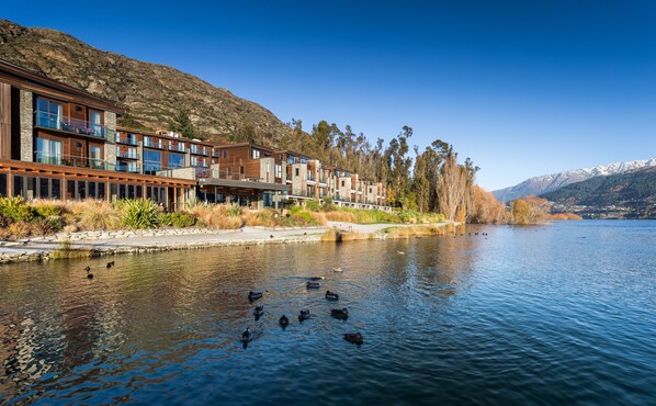 A lakeside hotel with modern architecture, a stone facade, and a view of the lake and mountains. - Hilton Queenstown Resort & Spa (Kawarau Falls)