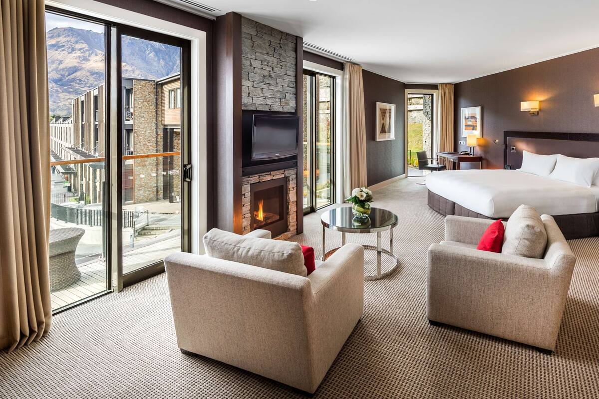 A hotel room with a large bed, two armchairs, a TV, a fireplace, and a balcony with a mountain view.