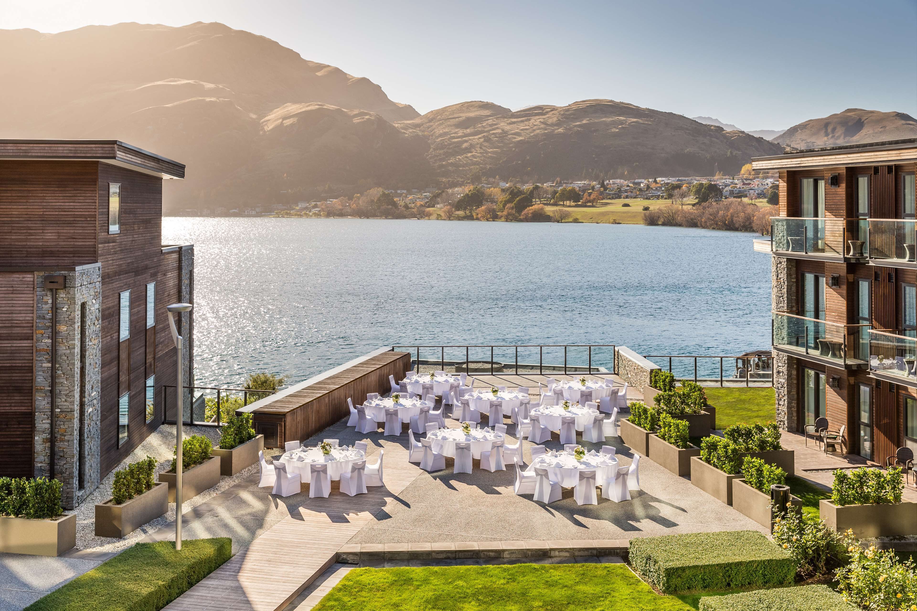 A lakeside outdoor event space with round tables set for an event, surrounded by modern buildings and a mountainous backdrop.