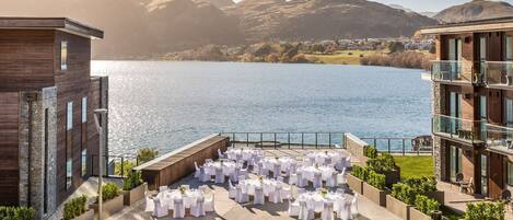 A lakeside outdoor event space with round tables set for an event, surrounded by modern buildings and a mountainous backdrop.