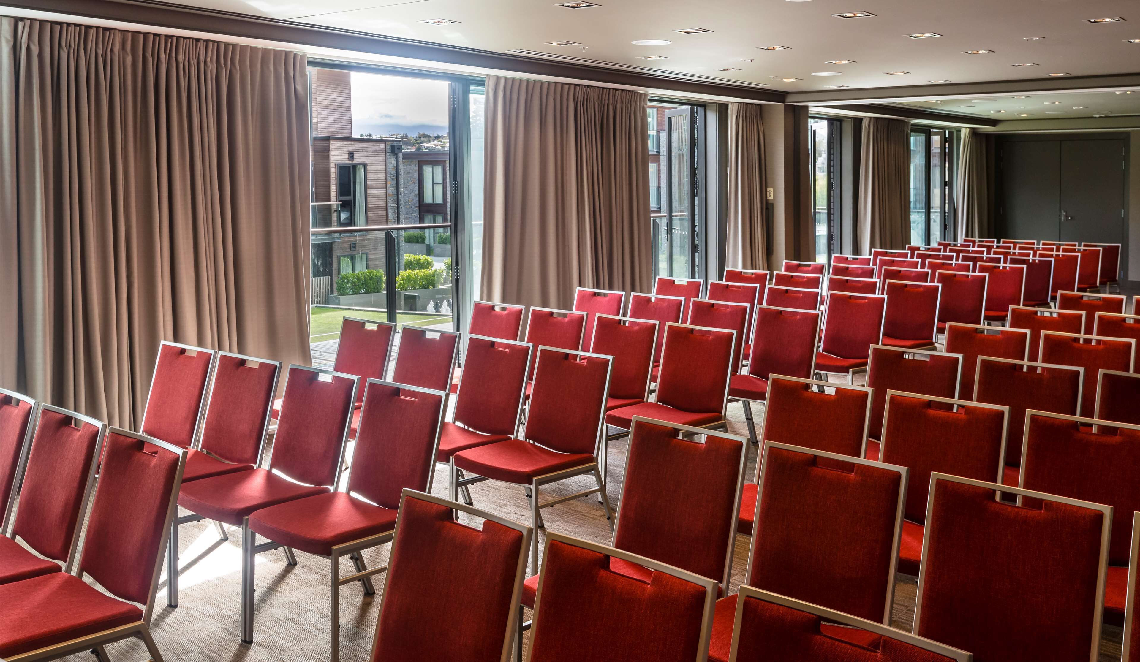 Een congreszaal met rijen rode stoelen, grote ramen voorzien van gordijnen en uitzicht op gebouwen.