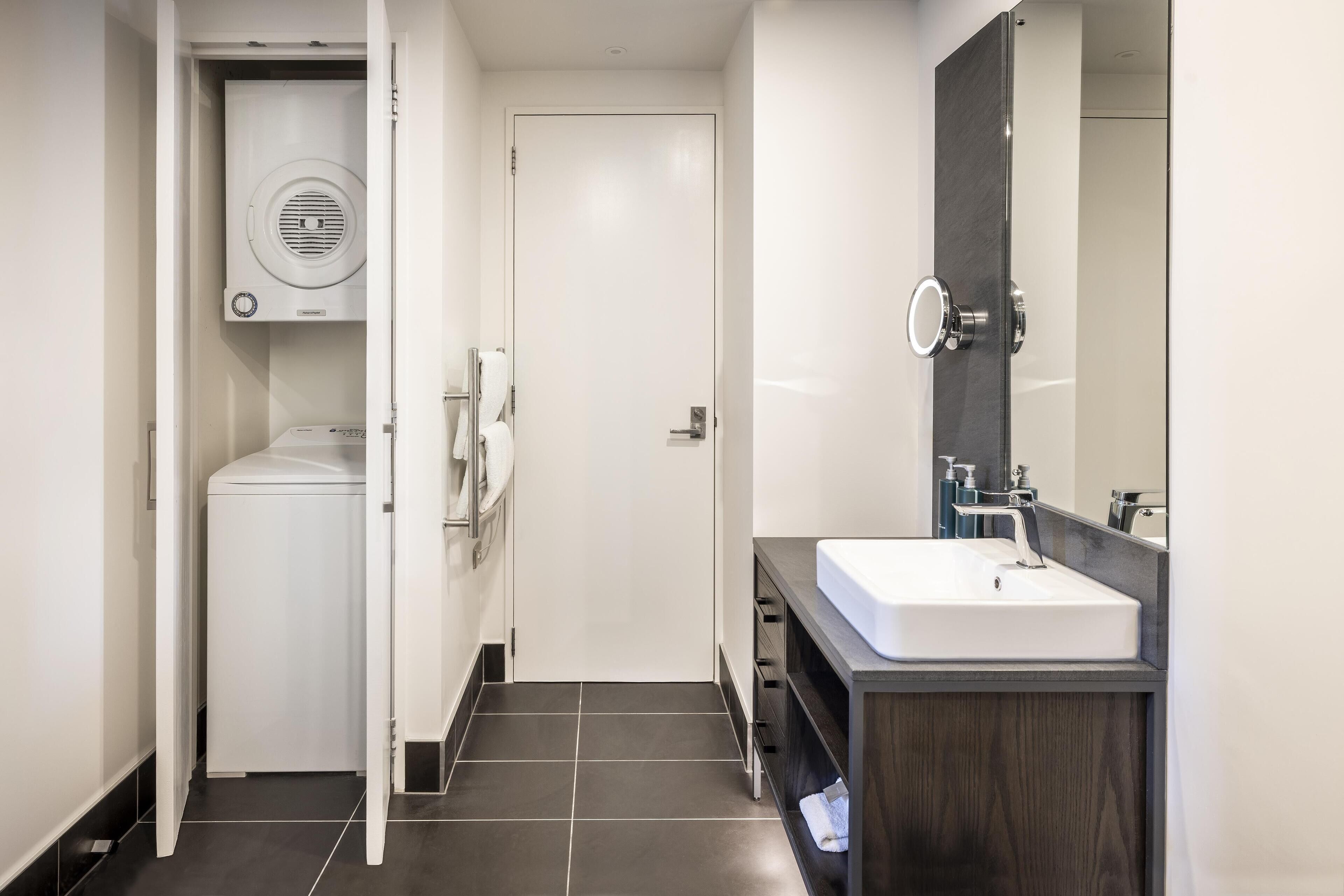 Un baño moderno con lavadora integrada, un lavabo con encimera oscura y un espejo sobre el lavabo.