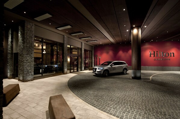 A modern hotel entrance with a stone pillar, a parked car, and a red wall with the hotel's name. - Hilton Queenstown Resort & Spa (Kawarau Falls)