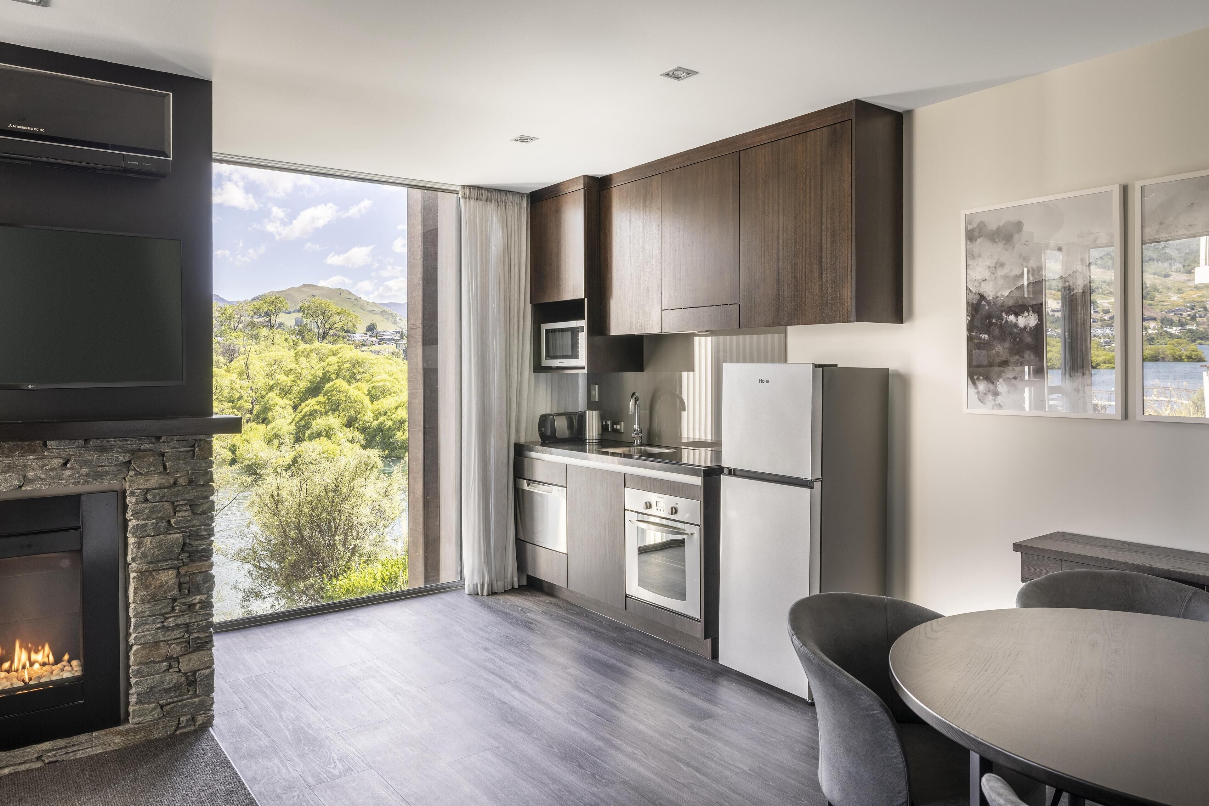 A modern kitchen with a stone fireplace, a dining area with a round table, and a view of greenery through large windows.