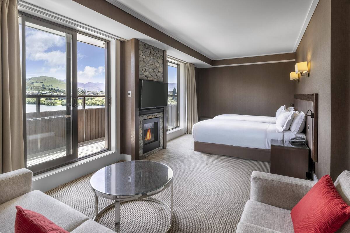 A hotel room with a large bed, a TV, a fireplace, and a balcony with a view of hills.