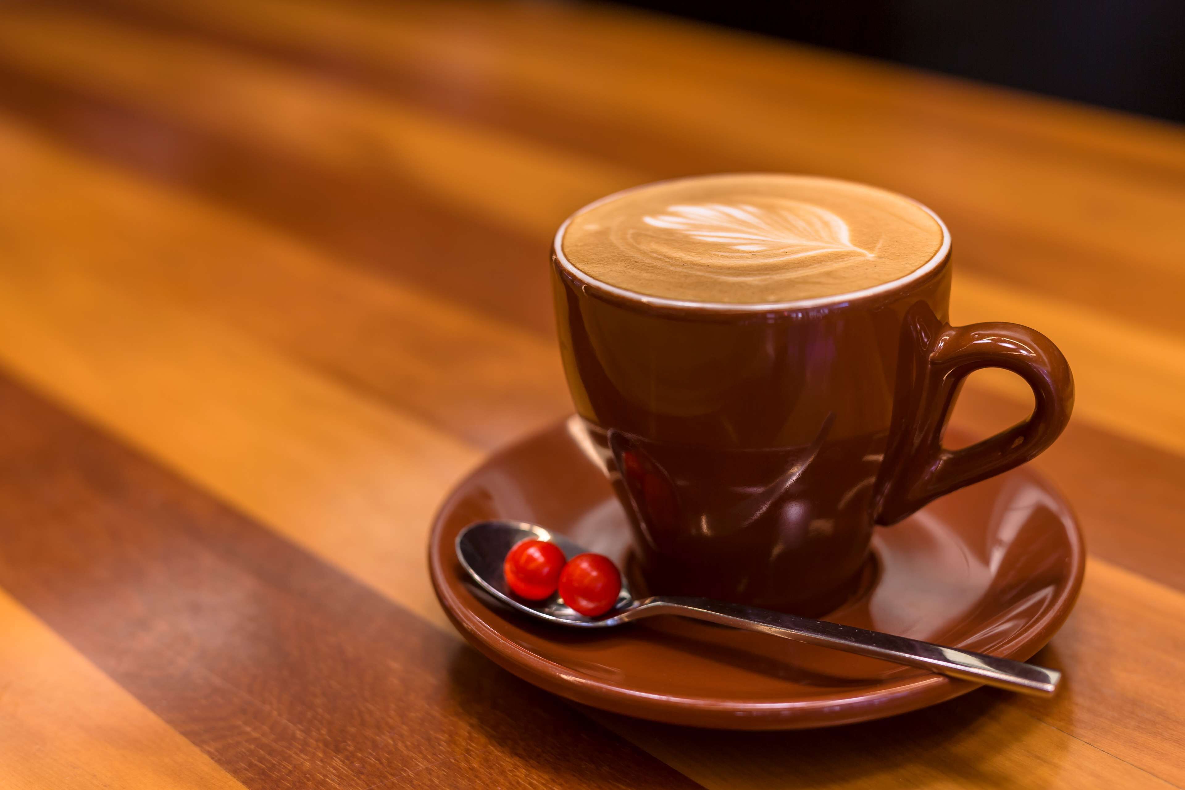 A cup of coffee with latte art on a saucer, accompanied by a spoon and two cherries.