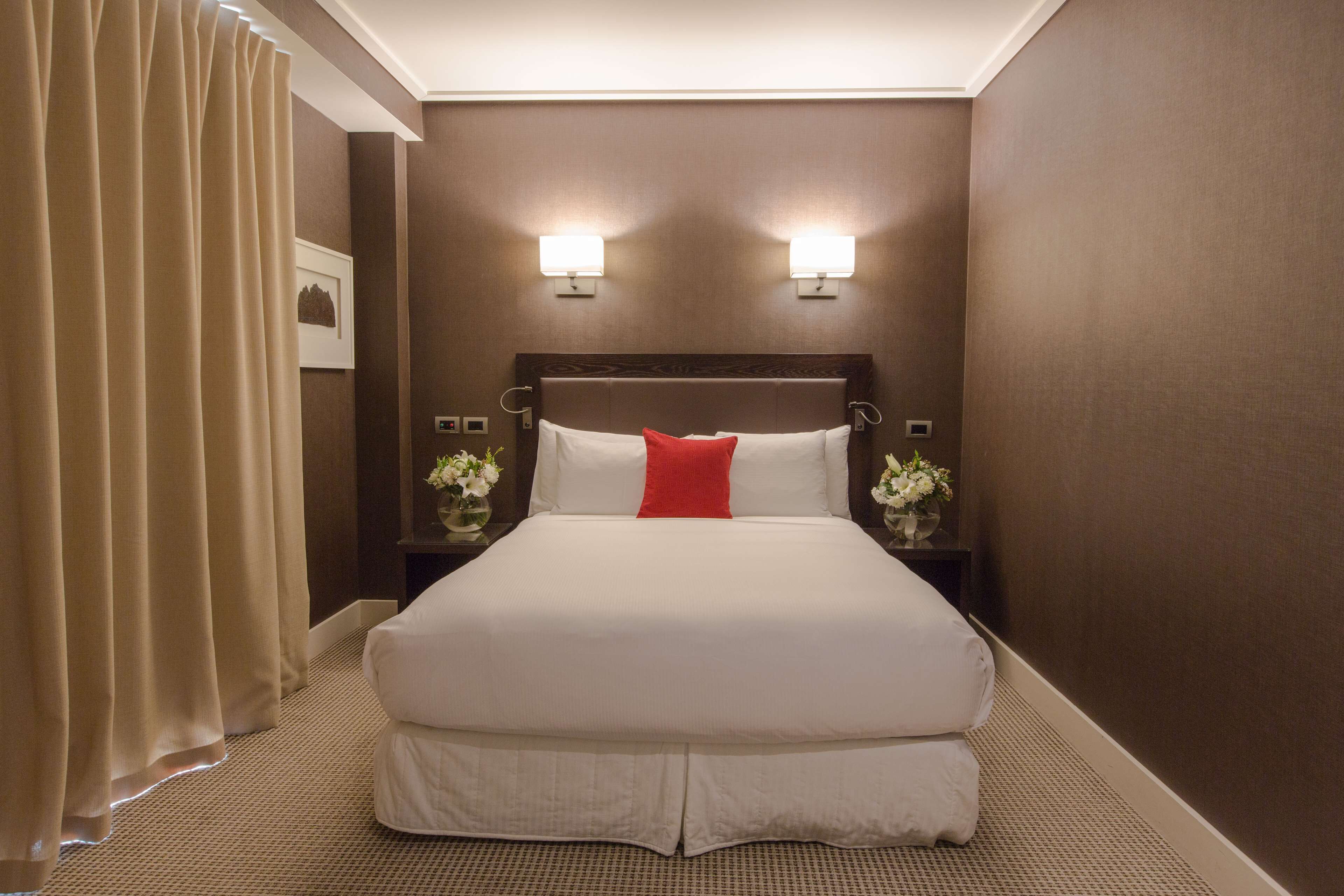 A hotel room with a large bed, two bedside lamps, a nightstand with a vase of flowers, and a curtain.