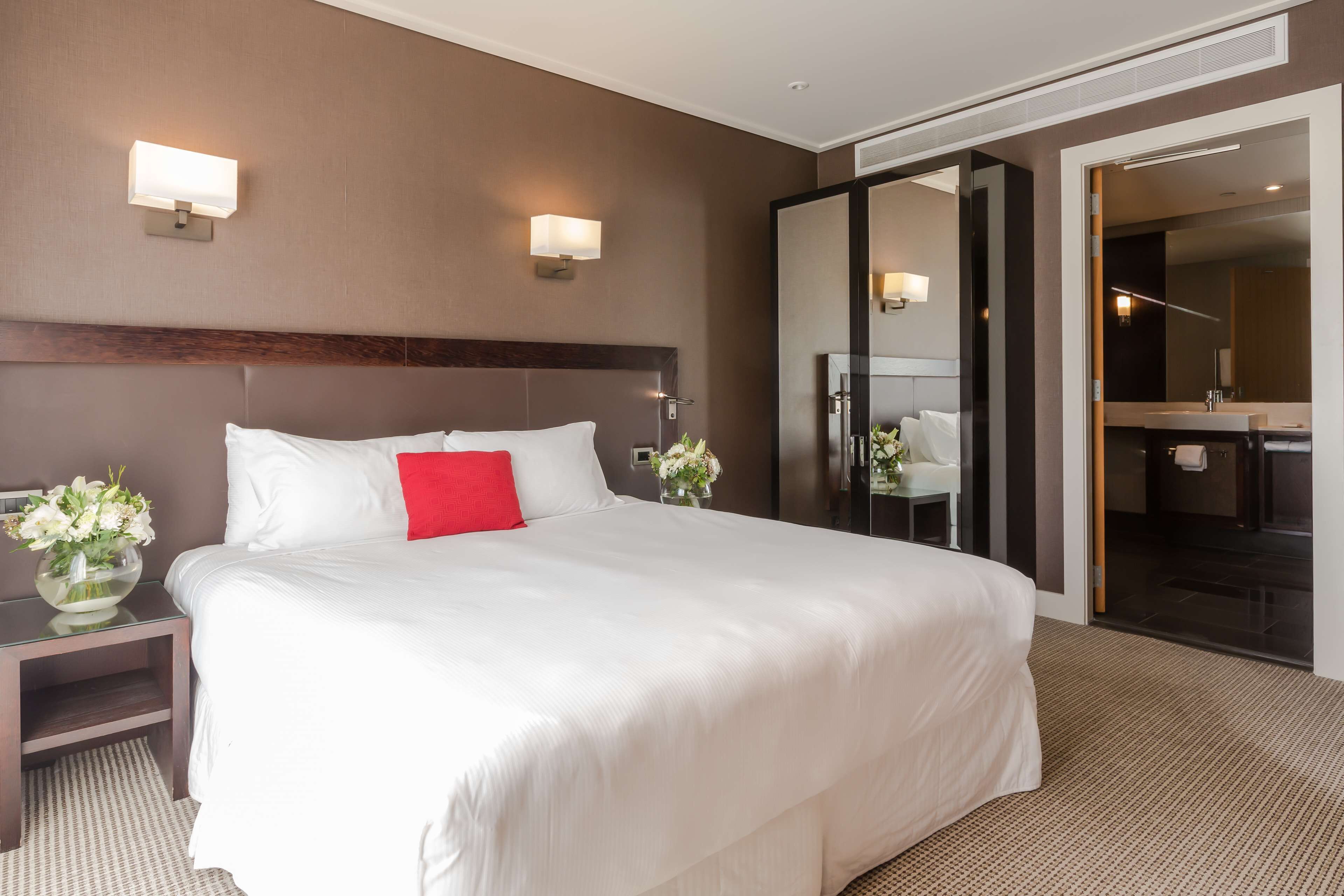 A hotel room with a large bed, a nightstand with a vase of flowers, a mirror, and a bathroom visible through an open door.