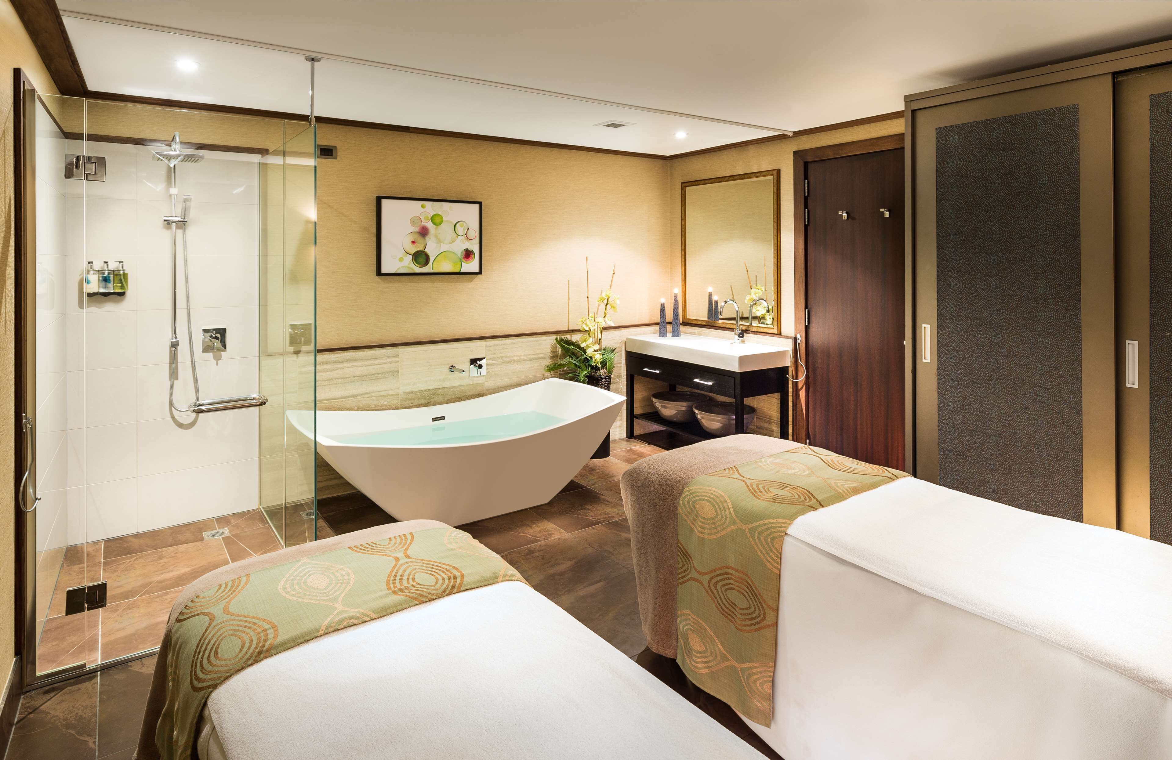 A modern hotel room with a glass-enclosed shower, a freestanding bathtub, a vanity with a mirror, and two beds with patterned bedspreads.