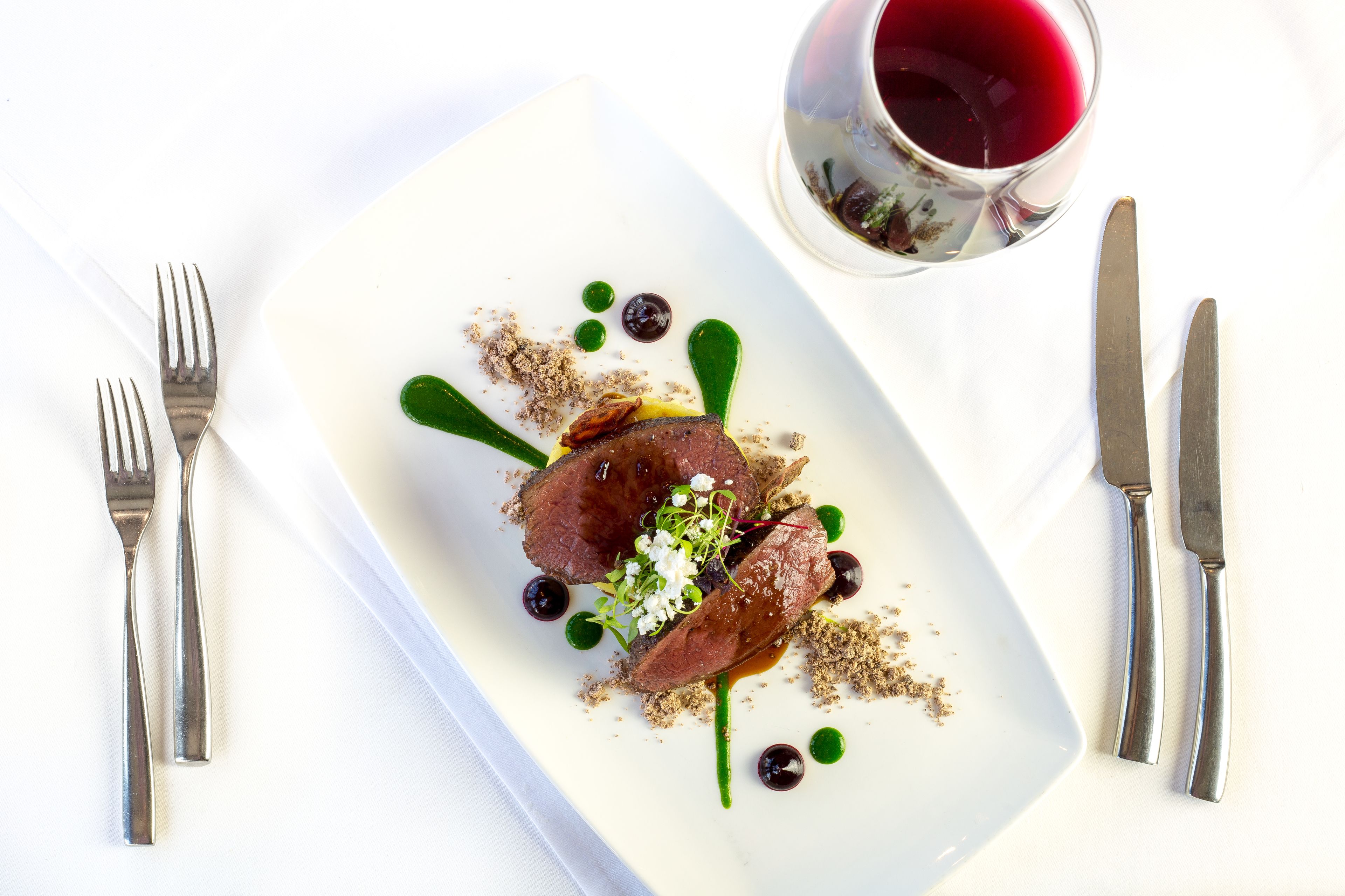A white rectangular plate with a piece of meat, garnishes, and a glass of red wine.