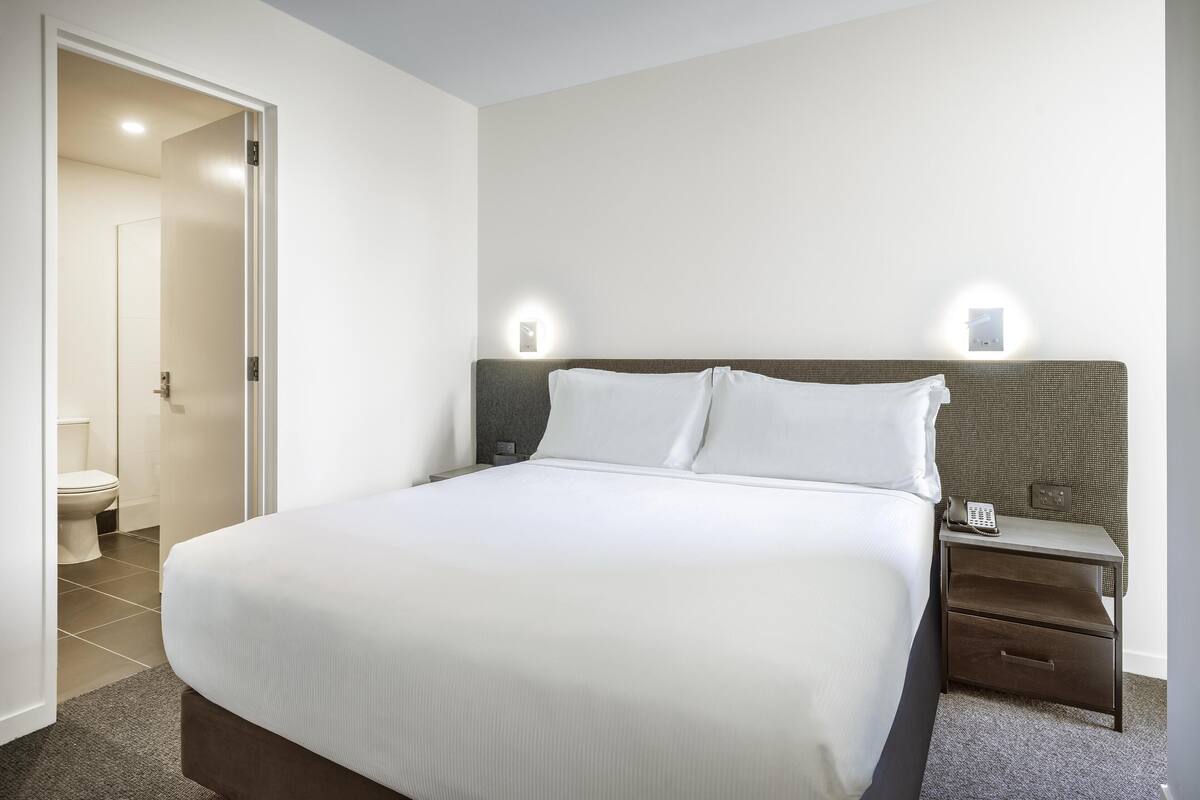 A neatly made bed with white linens, a nightstand with a lamp, and a wall-mounted light fixture.