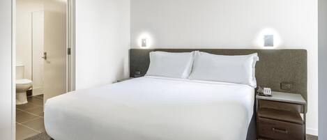 A neatly made bed with white linens, a nightstand with a lamp, and a wall-mounted light fixture.