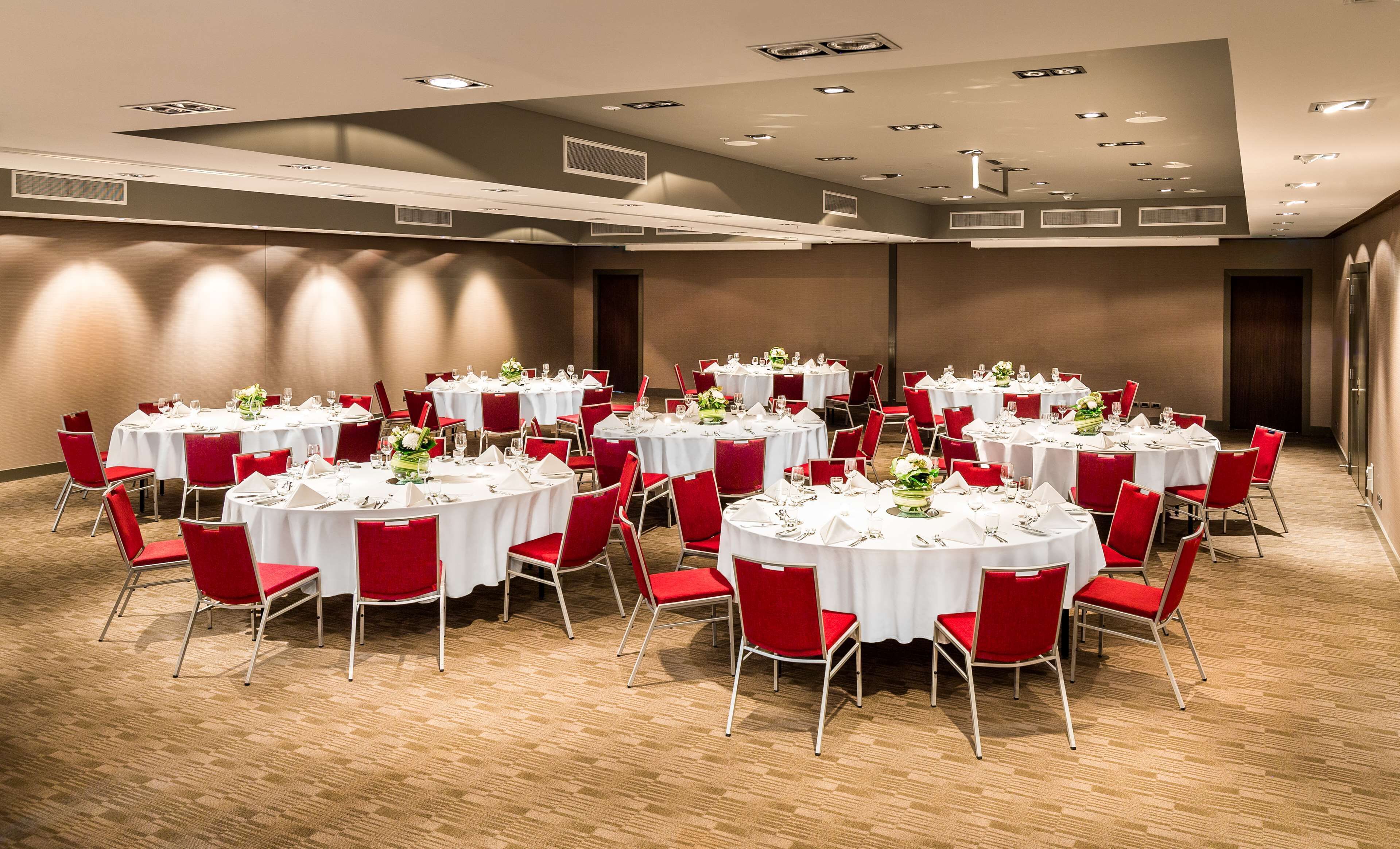 A conference room with multiple round tables, each set with white tablecloths, red chairs, and floral centerpieces.
