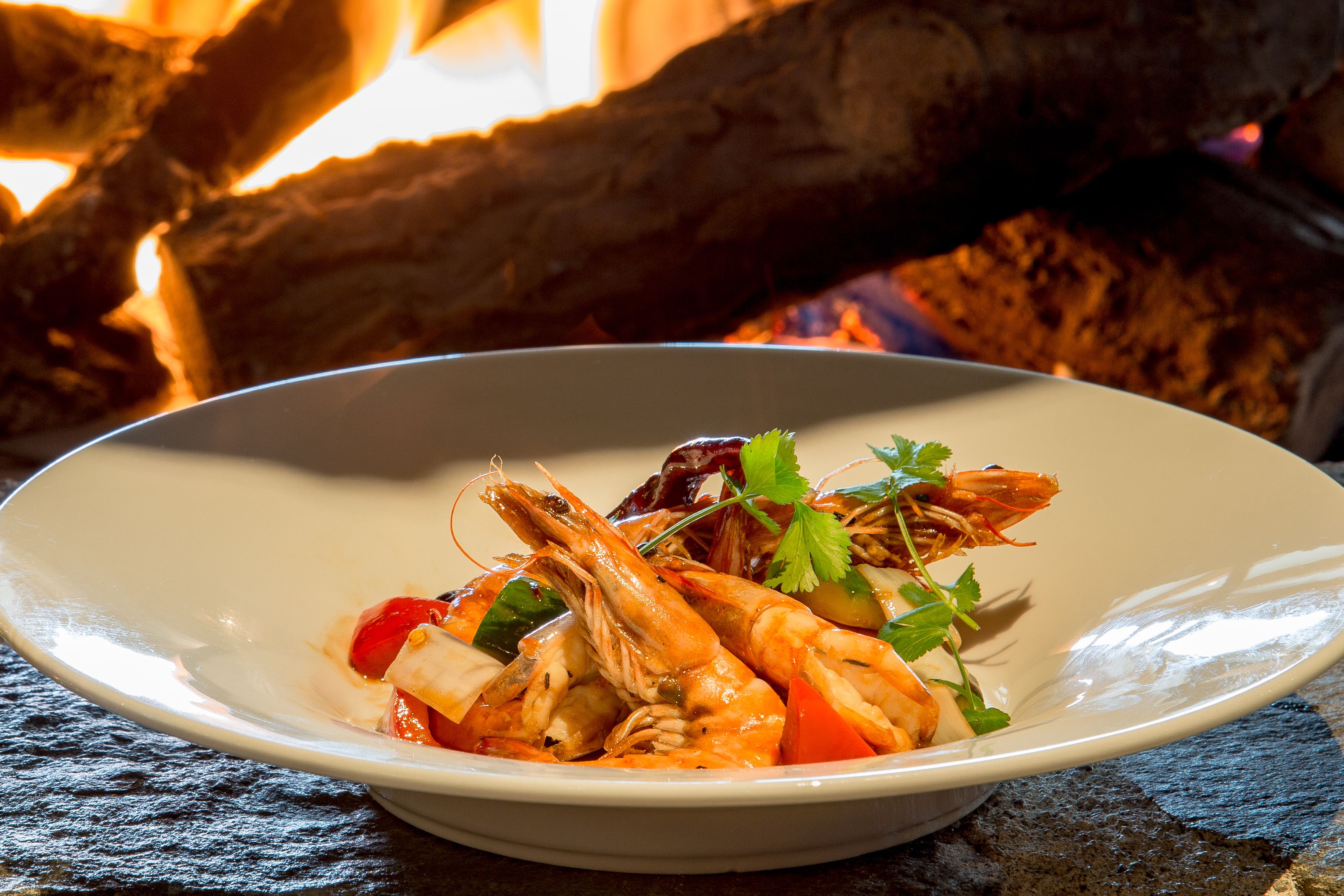 Un bol de crevettes et de légumes sur une surface en pierre, avec un feu en arrière-plan.