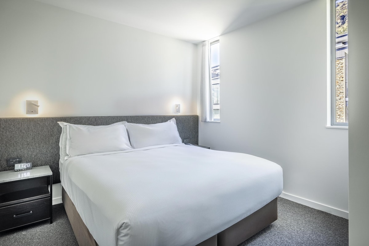 Un letto rifatto con cura in una camera d'albergo, con una testiera grigia, un comodino con una sveglia digitale e una finestra vista esterna.