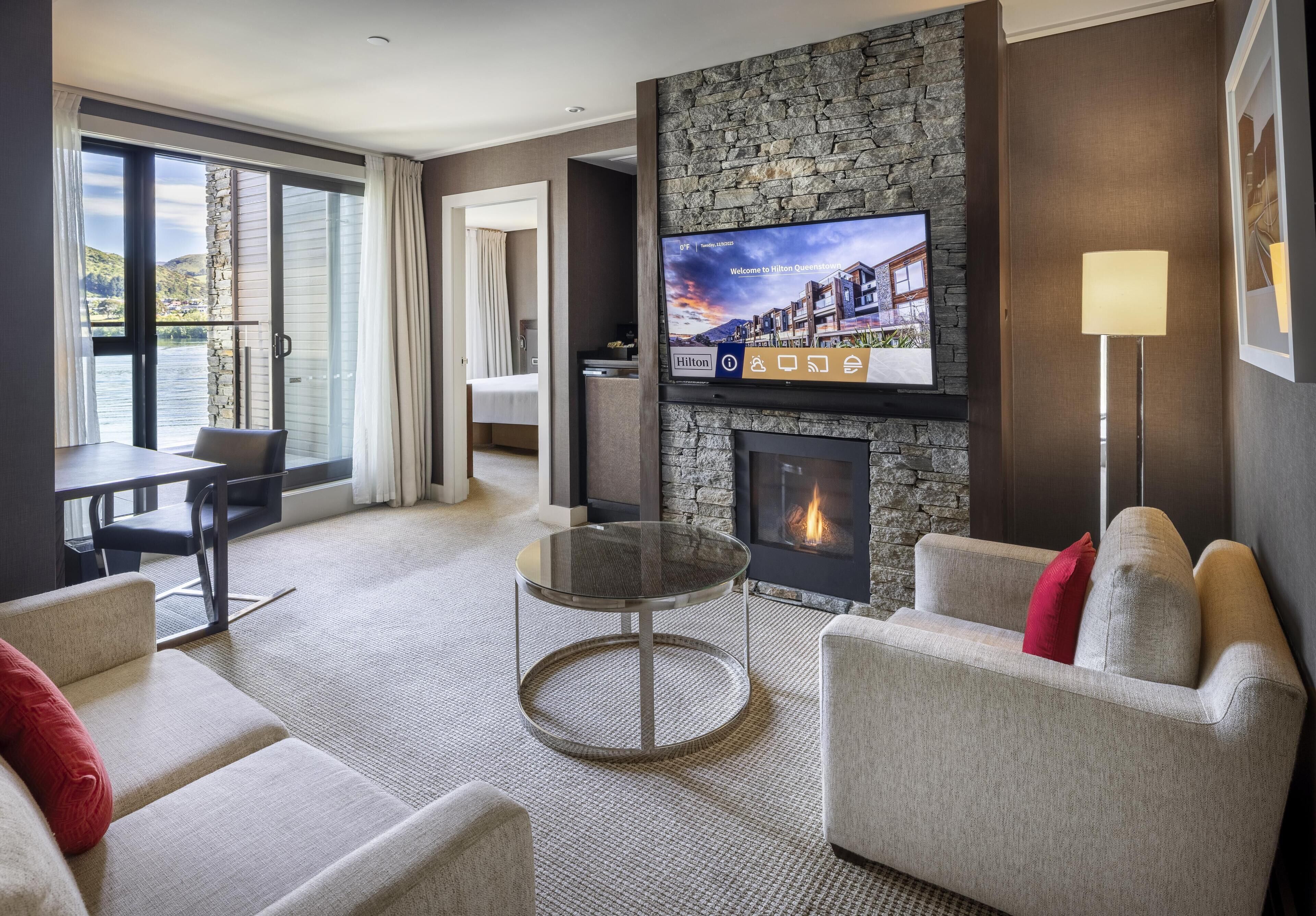 A hotel room with a fireplace, a television mounted on the wall, a dining area with a table and chairs, and a view of the outdoors through the windows.