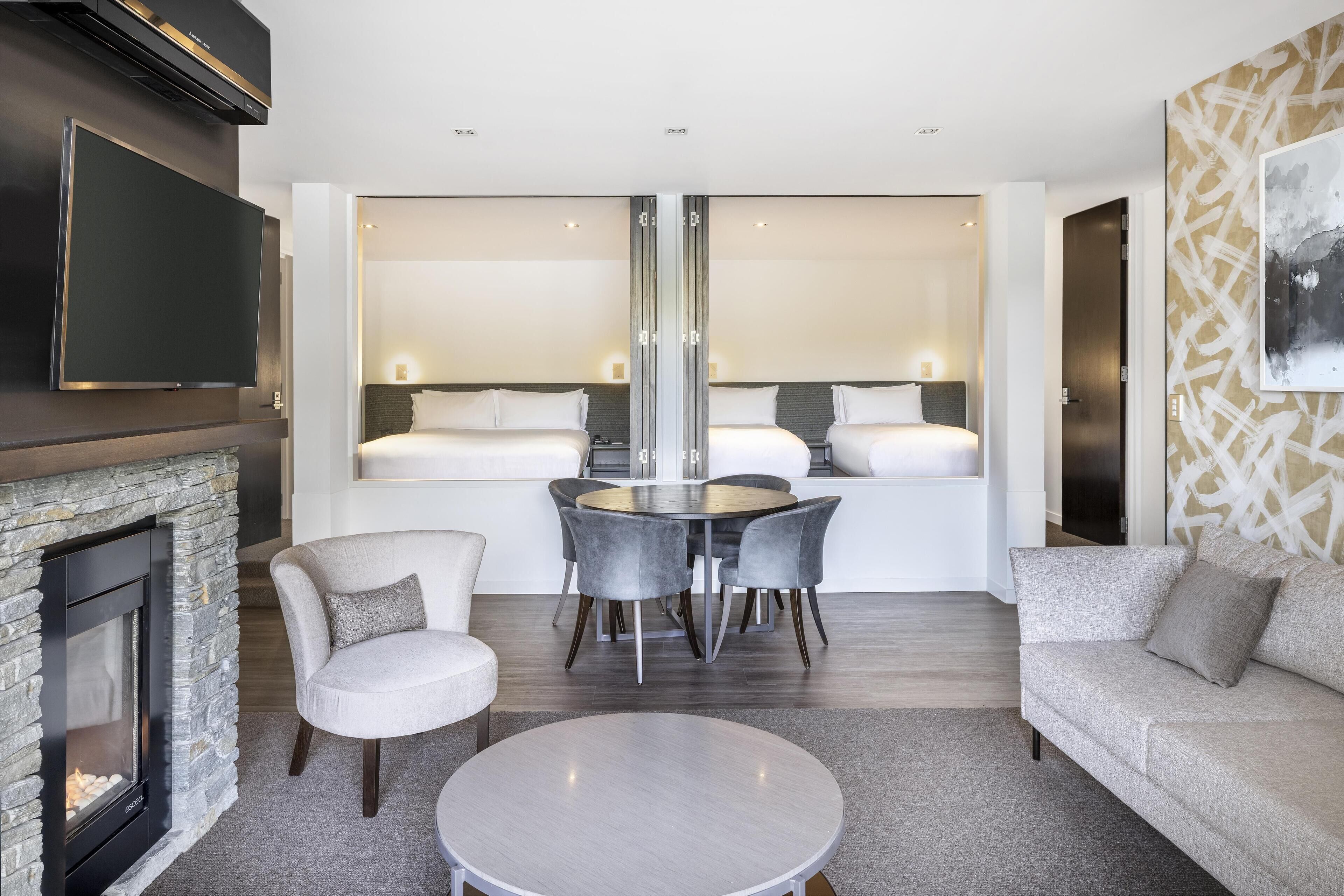 A modern hotel room with a fireplace, a TV mounted on the wall, a dining area with a round table and chairs, and a sofa.