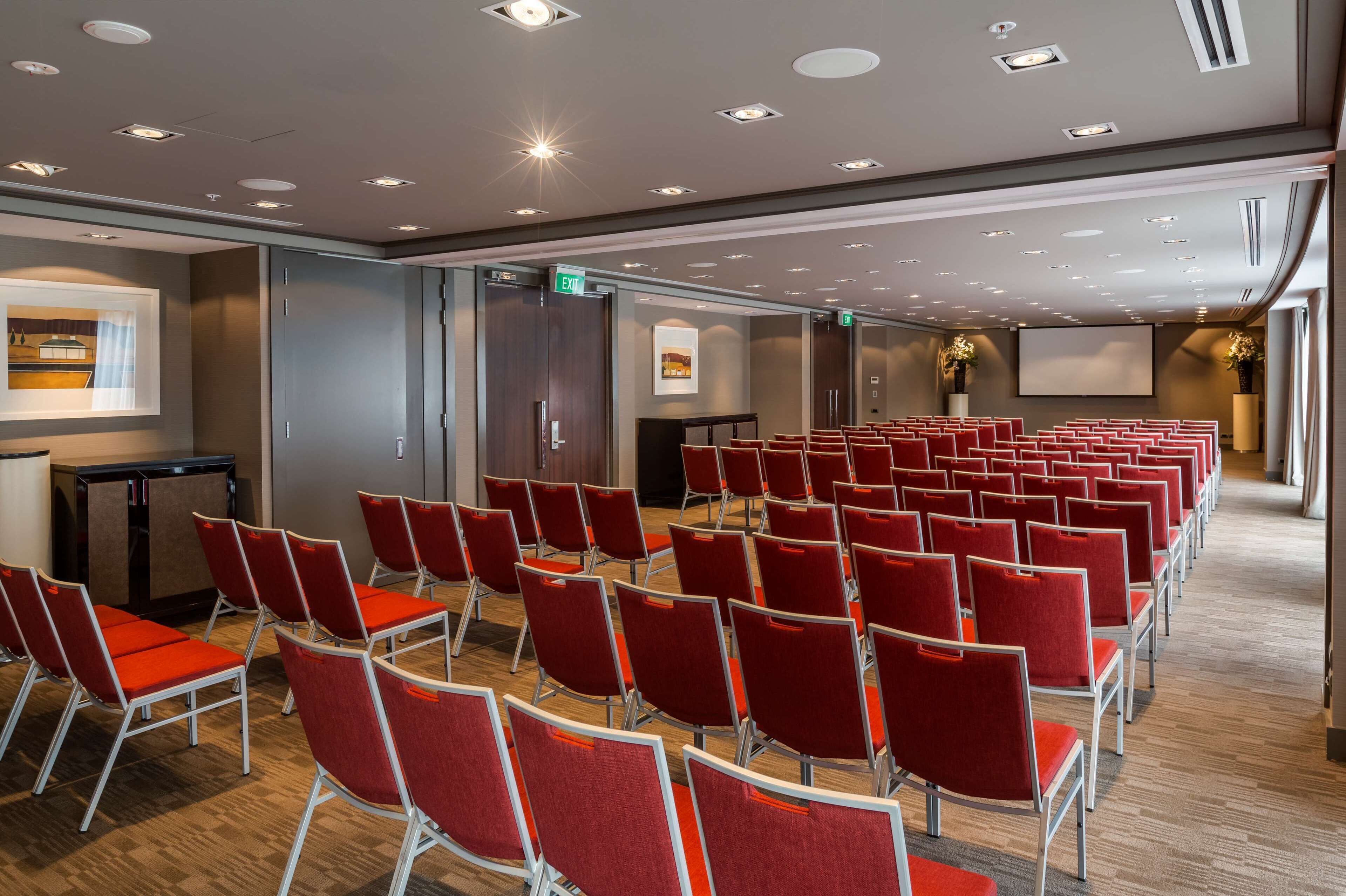 A conference room with rows of red chairs, a projector screen, and a podium.