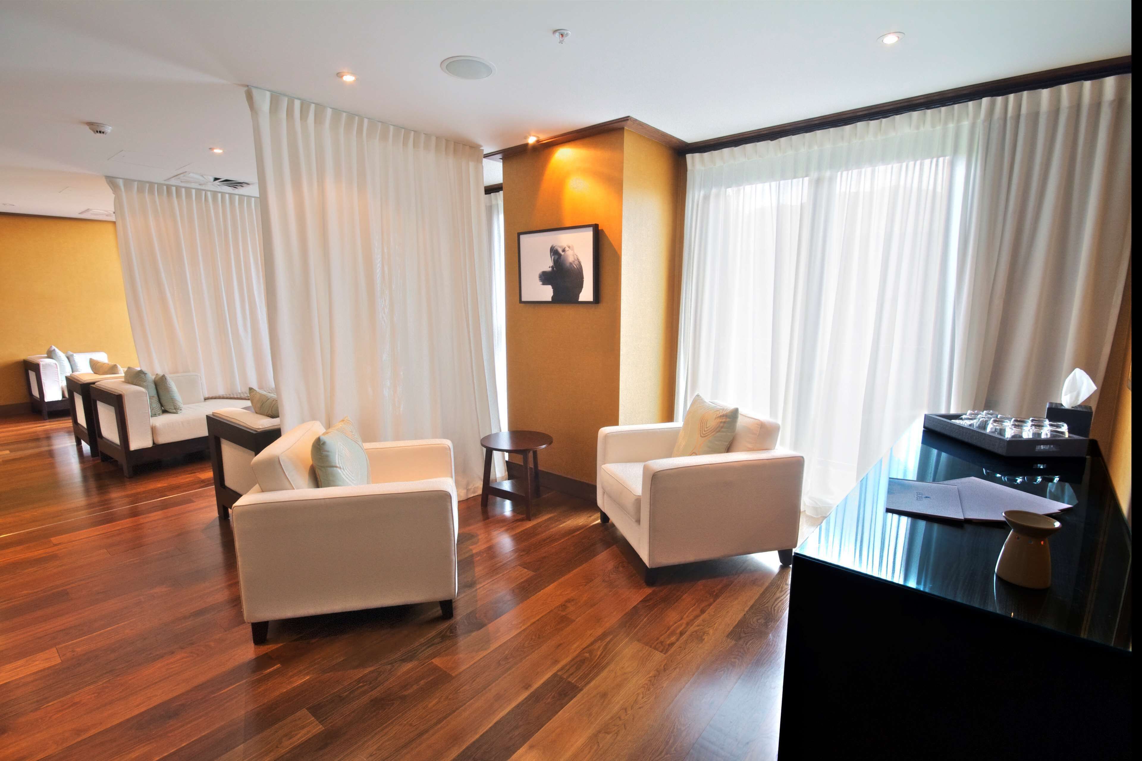 A modern hotel room with a wooden floor, two white sofas, a small round table, and a desk with a lamp and papers.