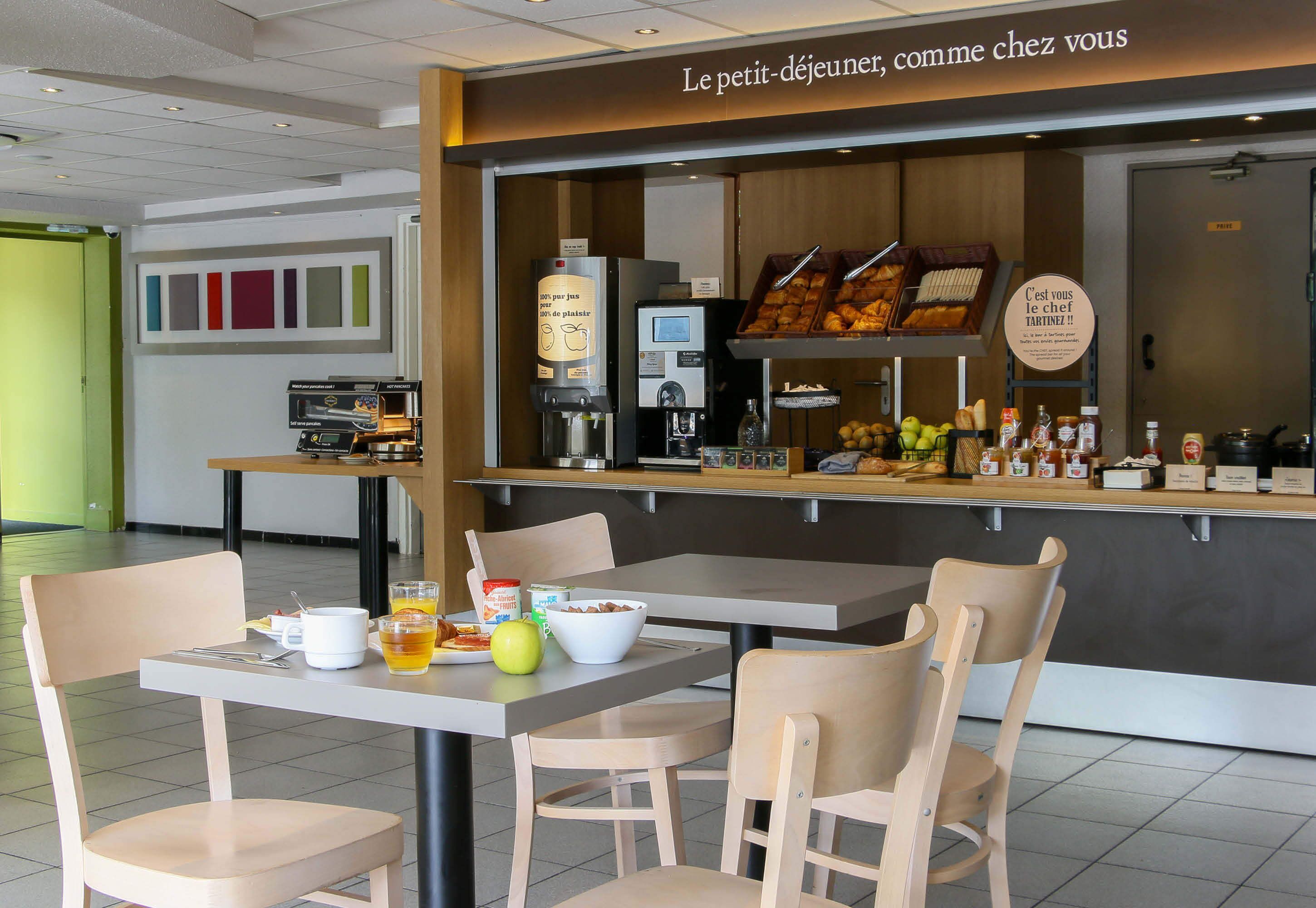 Ein Frühstücksbereich mit einer Kaffeemaschine, einer Obstauslage und einem Schild mit der Aufschrift 'Le petit-déjeuner, comme chez vous'.