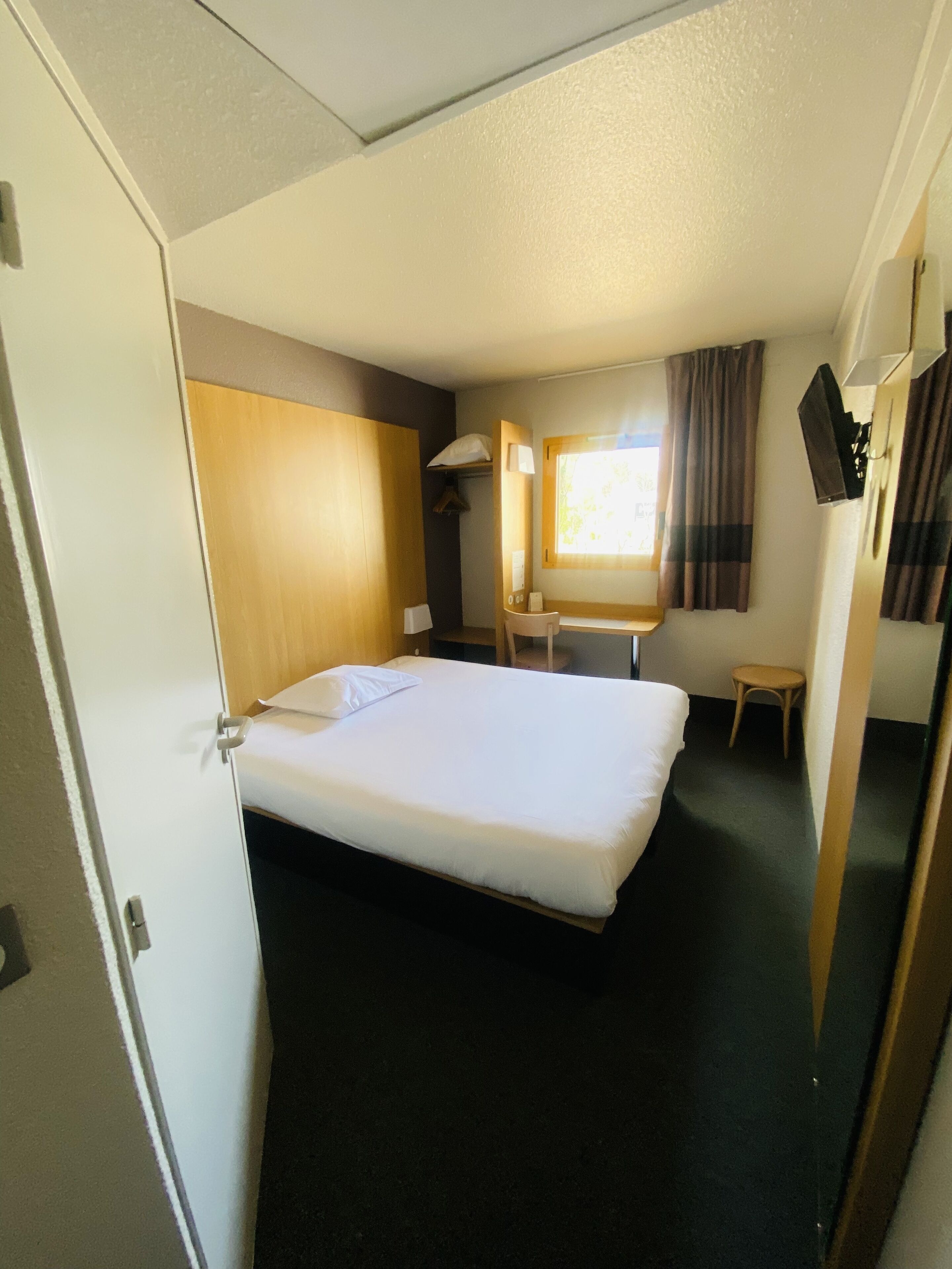 Ein Hotelzimmer mit einem Bett, einem Schreibtisch, einem Stuhl, einem kleinen Tisch, einem Spiegel und einem Fenster mit Vorhängen.