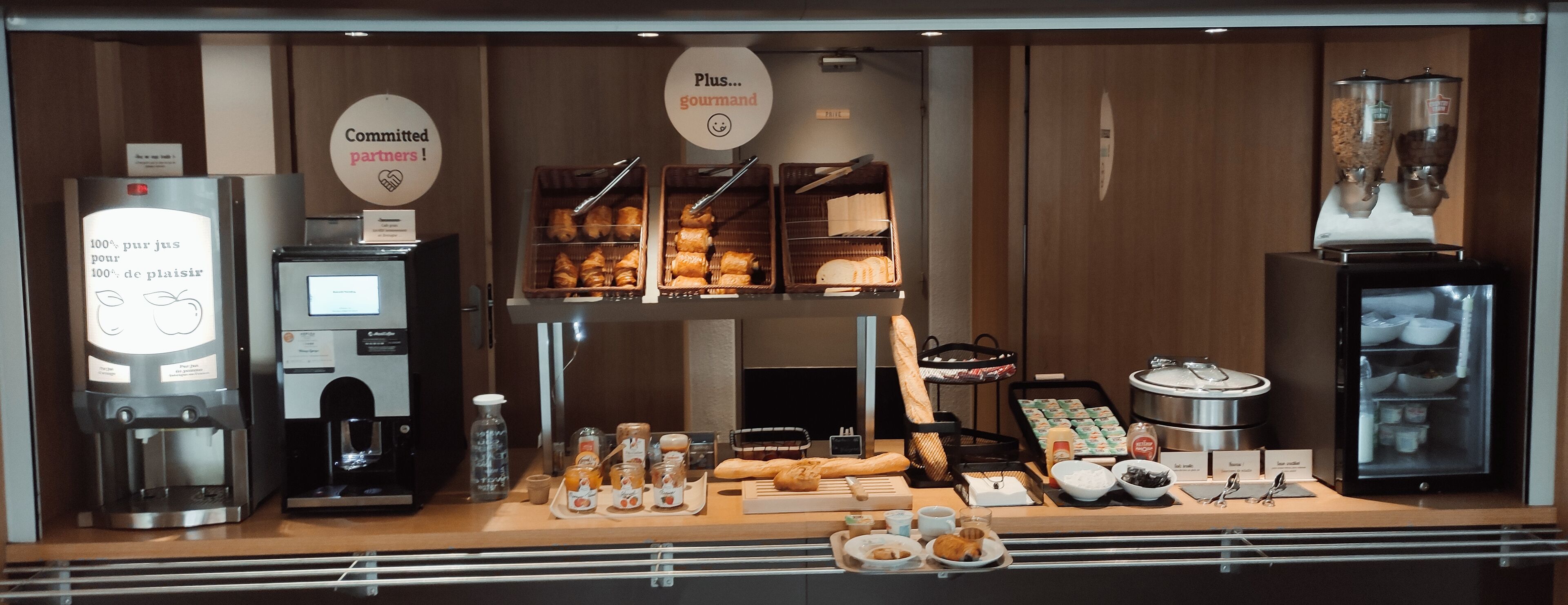 A breakfast buffet with various food items, coffee machines, and a refrigerated display.