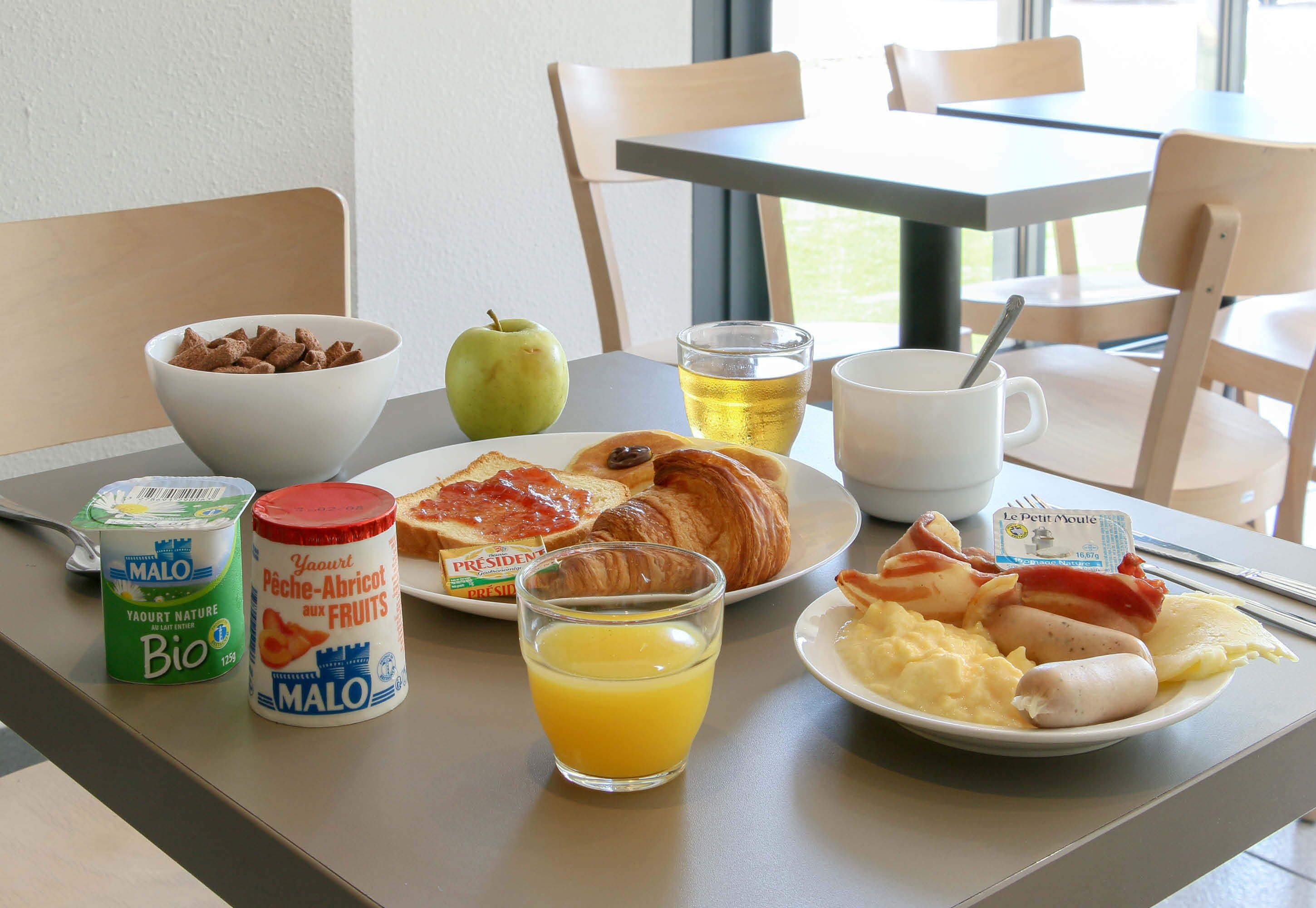 A breakfast table with a bowl of cereal, a glass of orange juice, a croissant, a plate with eggs and bacon, a glass of tea, and a container of yogurt.