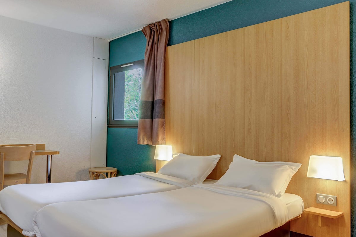 A hotel room with two beds, a wooden headboard, a small table, a chair, and two bedside lamps.