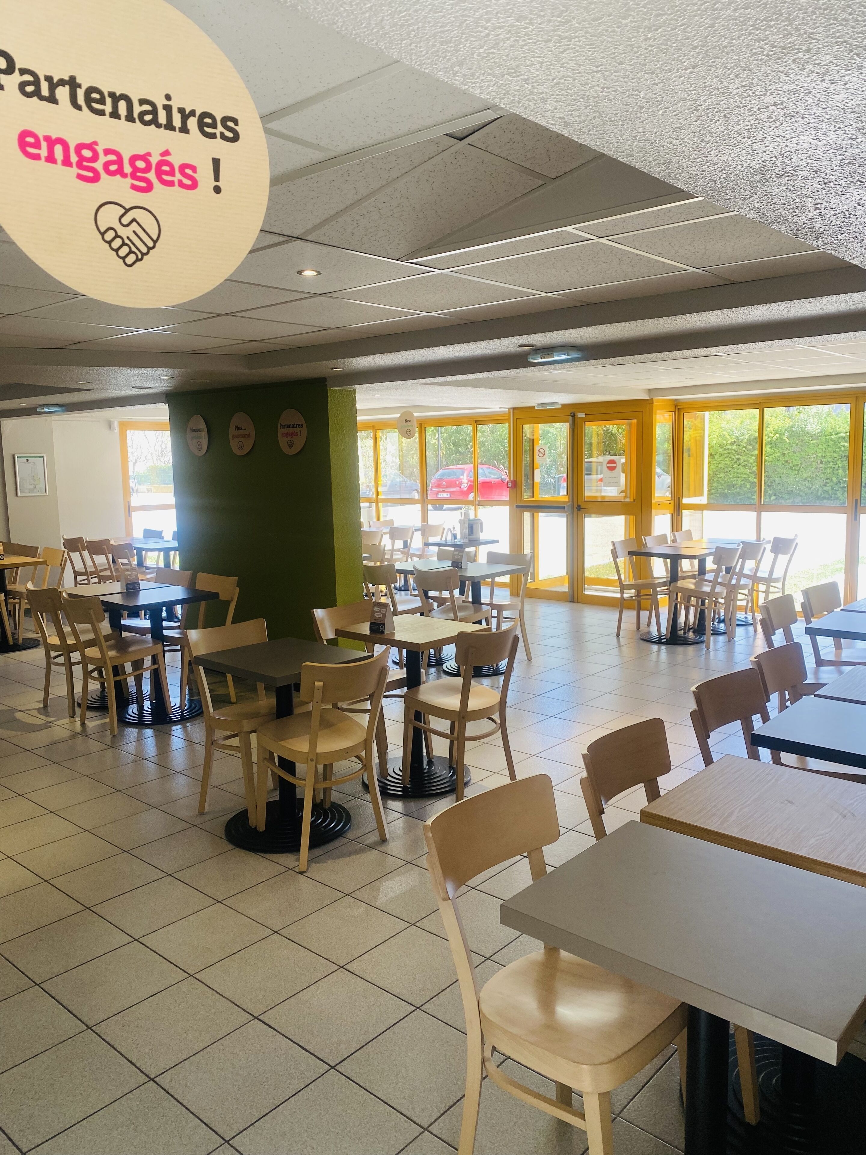 A cafeteria with tables and chairs, a sign reading "Partenaires engagés !", and large windows allowing natural light.