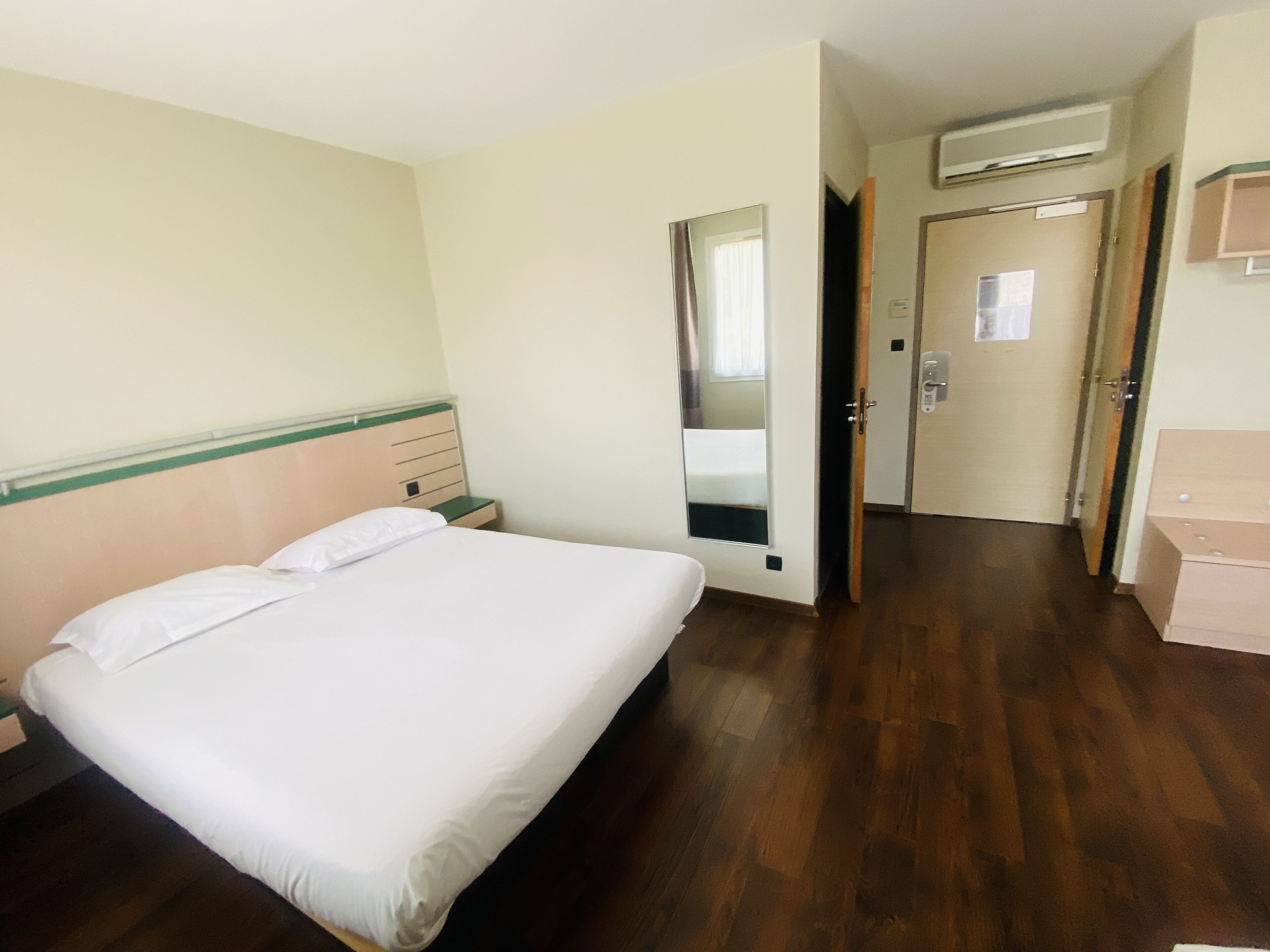 A hotel room with a single bed, a wooden floor, a mirror, a door, and a small bedside table.