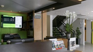 Una recepción con un mostrador y una pantalla digital que muestra tarifas, una escalera y plantas en macetas.