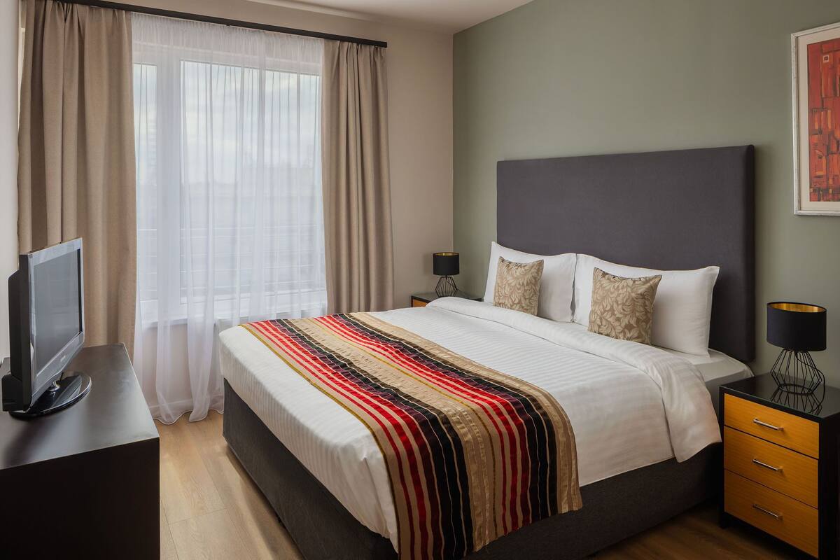 A hotel room with a large bed, a TV on a stand, a bedside table with a lamp, and a window with sheer curtains.