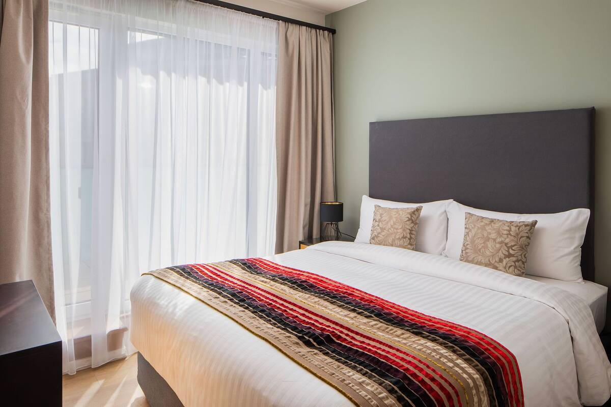 A hotel room with a large bed, a headboard, a bedside table, and a window with sheer curtains.