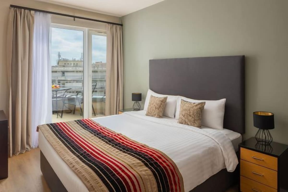 A hotel room with a large bed, a nightstand, a lamp, and a view of the city through a window.
