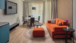 A modern living room with a sofa, ottoman, and a small table with a plant. There is a dining area with chairs and a table, and a TV mounted on the wall. - Escala Hotel & Suites (Budapest)