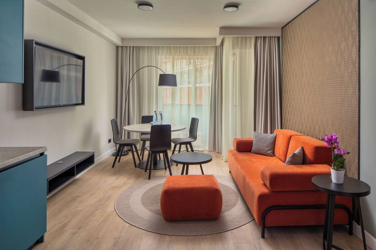 A modern living room with a sofa, ottoman, and a small table with a plant. There is a dining area with chairs and a table, and a TV mounted on the wall.