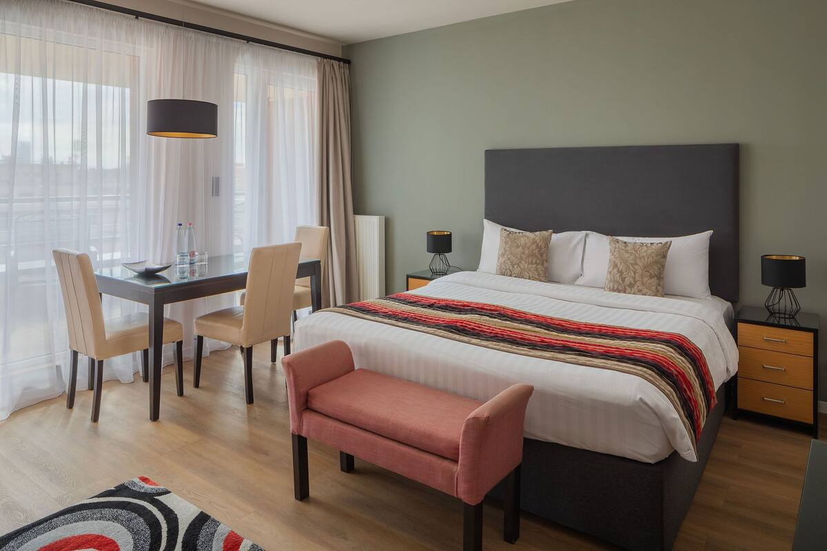 A modern hotel room with a large bed, bedside tables, a desk with chairs, and a view through sheer curtains.