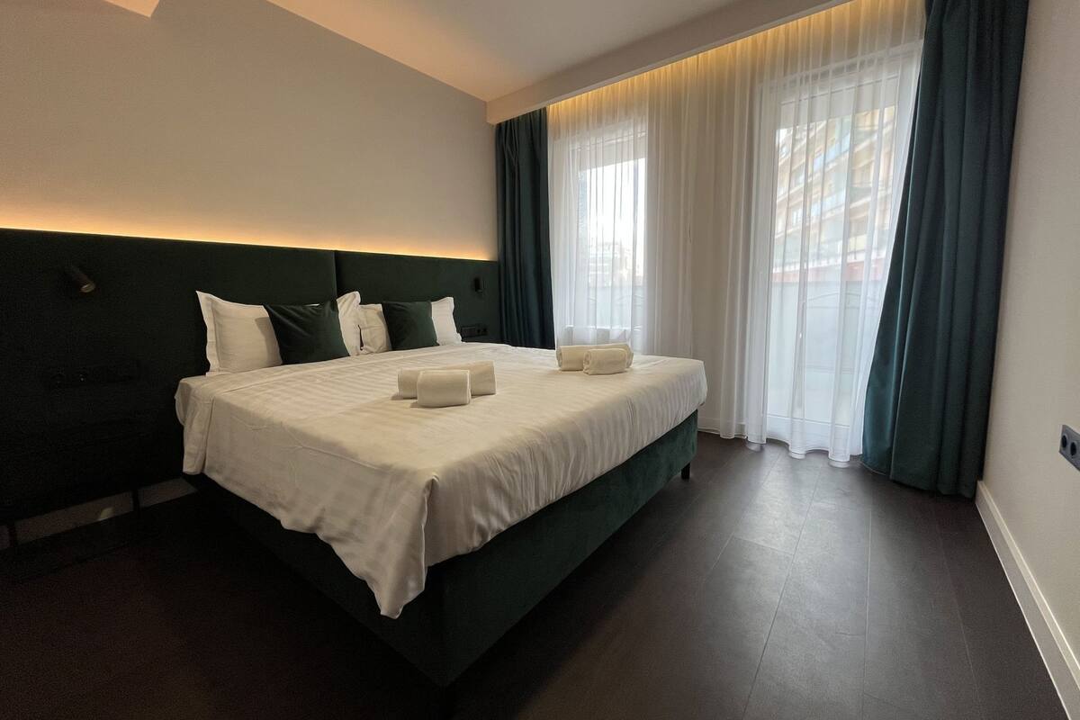 A hotel room with a large bed, dark green headboard, white bedding, and sheer curtains.