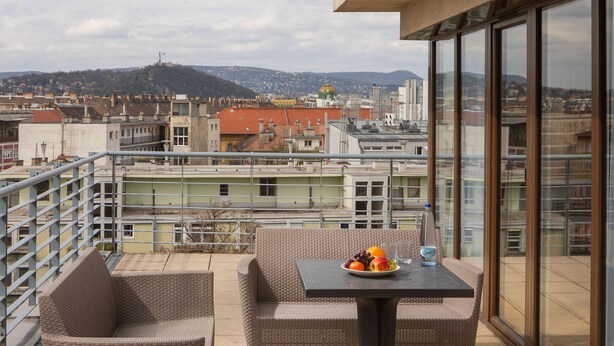A balcony with wicker furniture, a table with fruit and a bottle, and a city view with buildings and a hill. - Escala Hotel & Suites (Budapest)