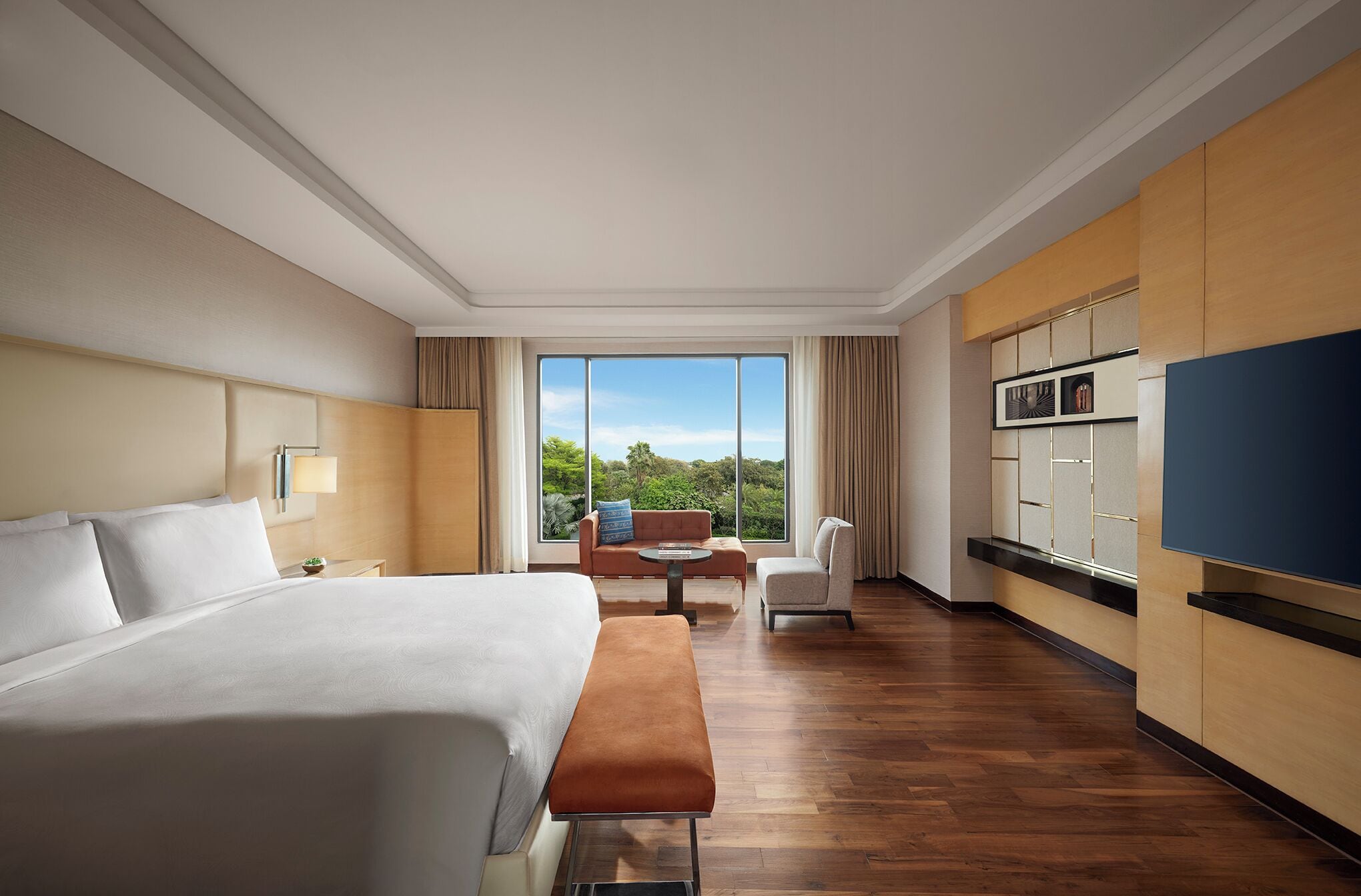 A hotel room with a large bed, a bench, a dining table with chairs, a TV, and a view of greenery outside.