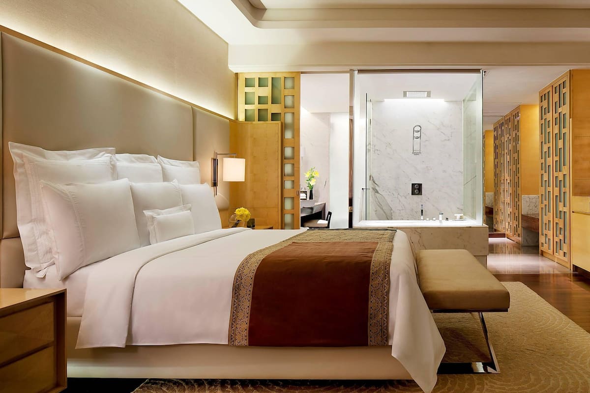 A modern hotel room with a large bed, a bedside table, a bench, a mirror, a vanity, and a shower area.