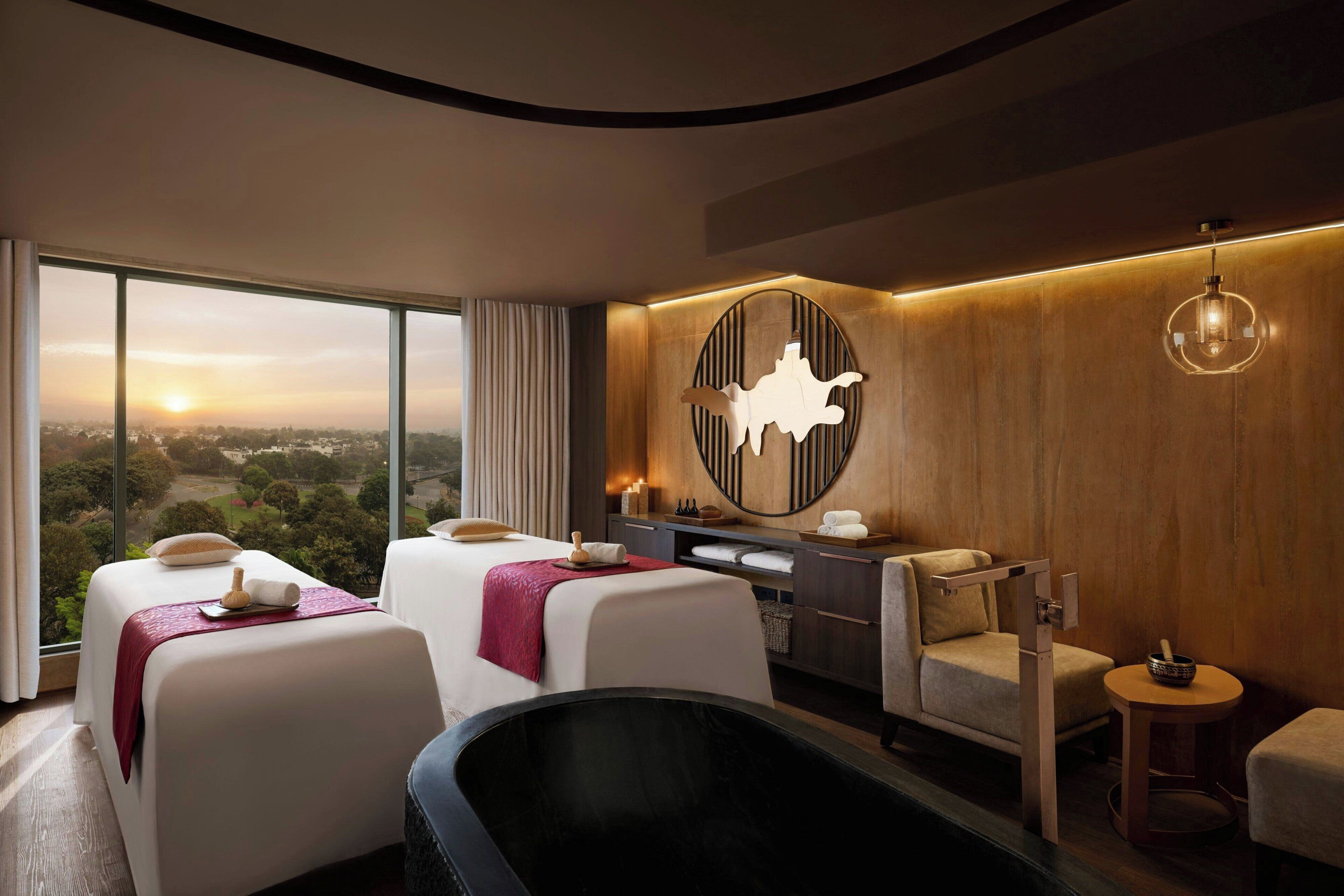 A modern hotel room with a large window, a massage table, a bathtub, and a wooden wall panel.