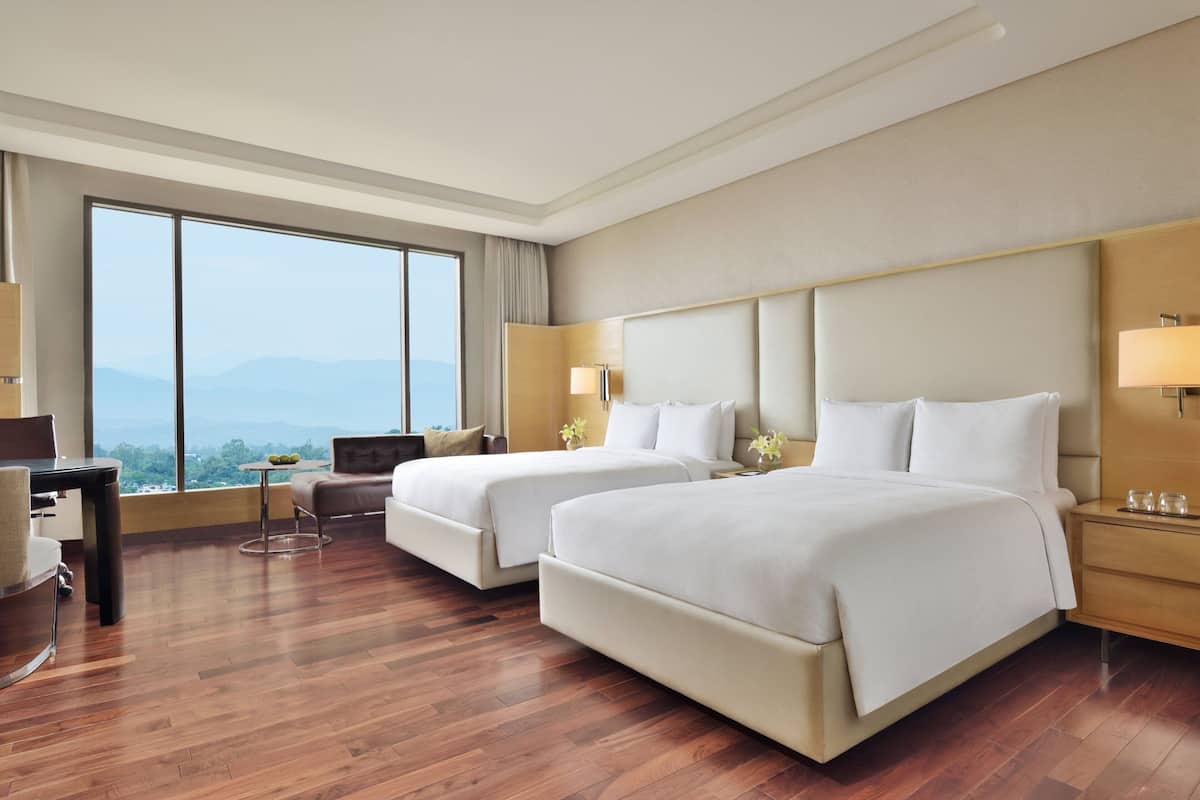 A hotel room with two beds, a desk, and a large window offering a mountain view.