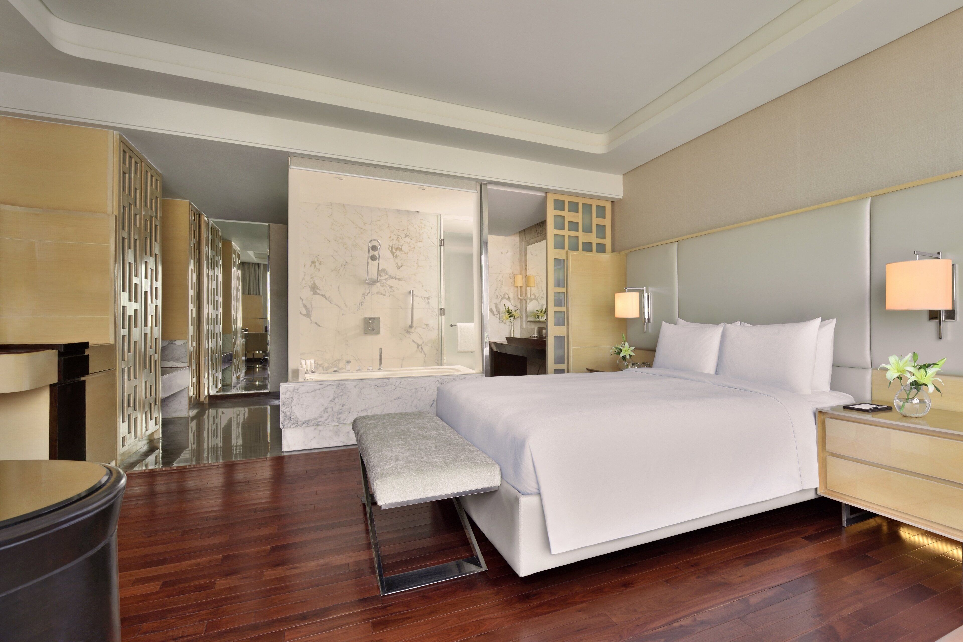 A modern hotel room with a large bed, a bench, a nightstand, a lamp, and a view of the bathroom.
